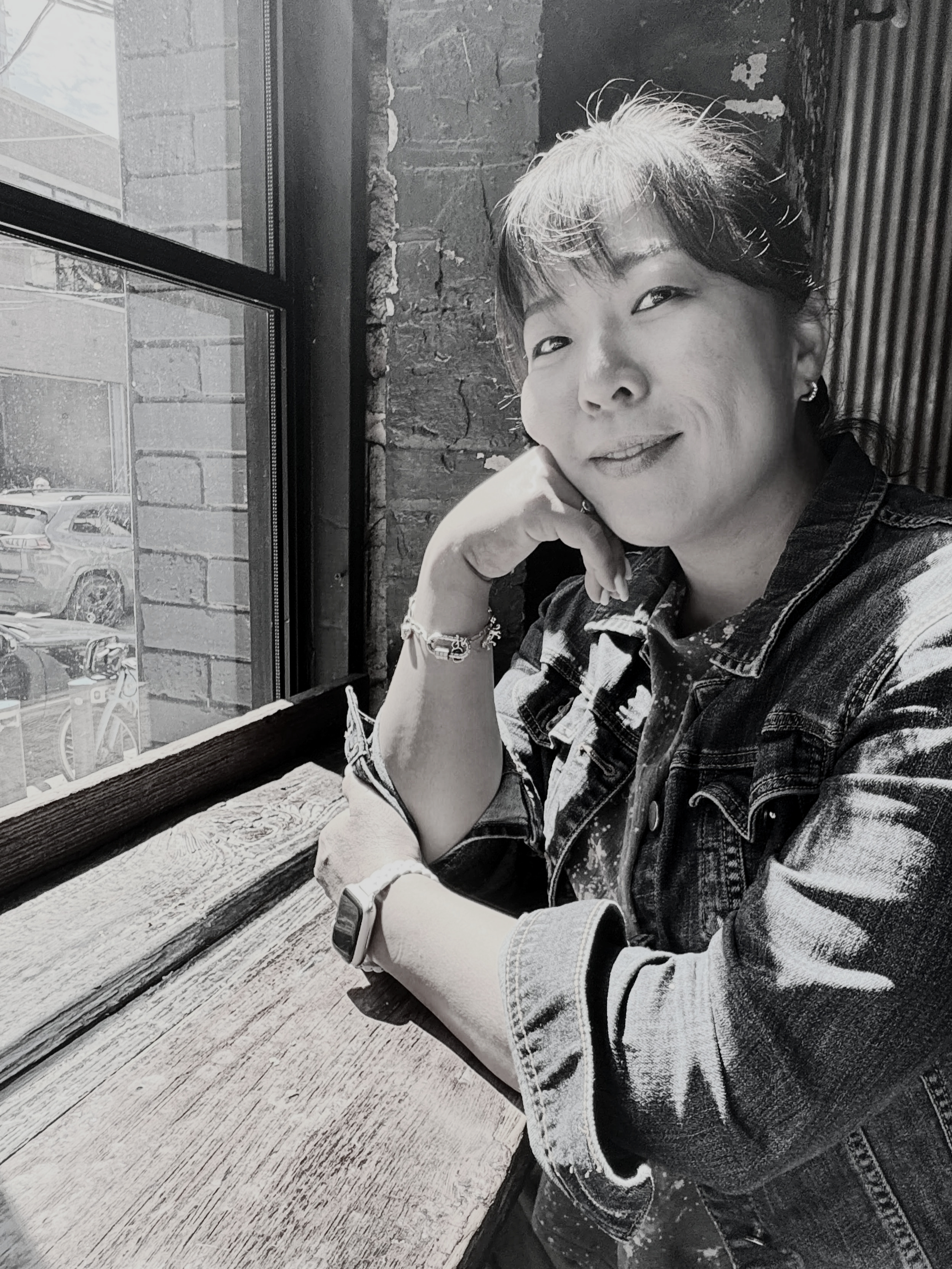 A woman with short hair wearing a denim jacket and a watch, sitting by a window with her head resting on her hand, smiling at the camera.