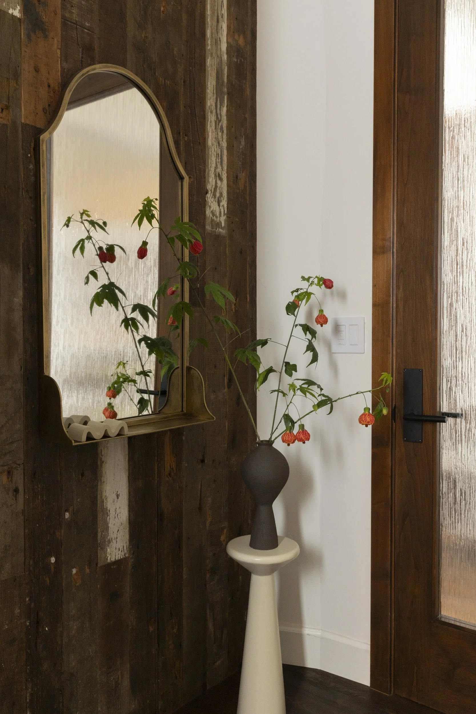 A reflection of a plant with pink flowers in a mirror mounted on a wooden wall next to a white pedestal with a dark vase and pink flowers. The scene is indoors with a white wall and wooden door.