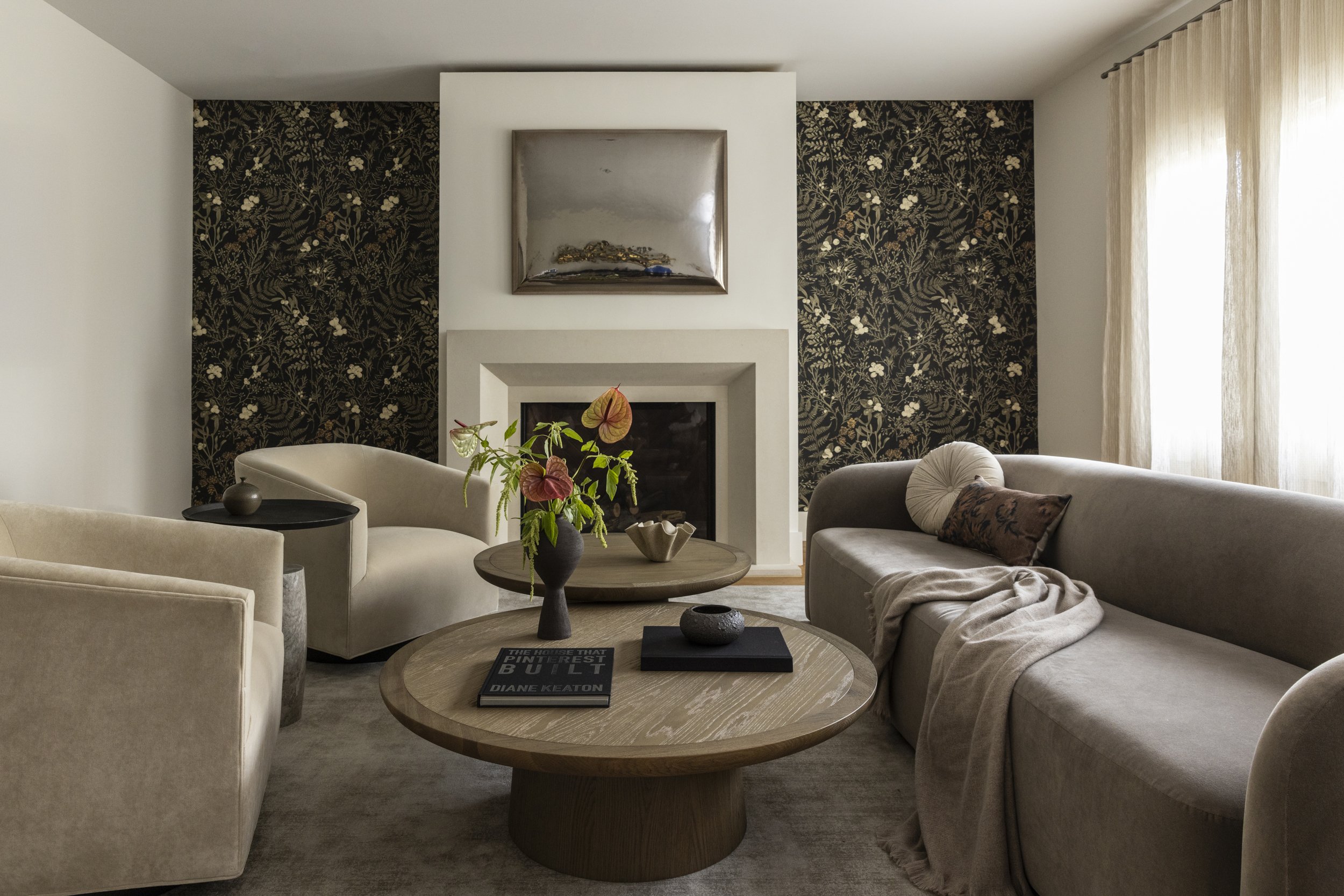 Living room with beige sofas, a round wooden coffee table with decorative objects and a plant, a fireplace with a mirror, and dark floral wallpaper on the walls.