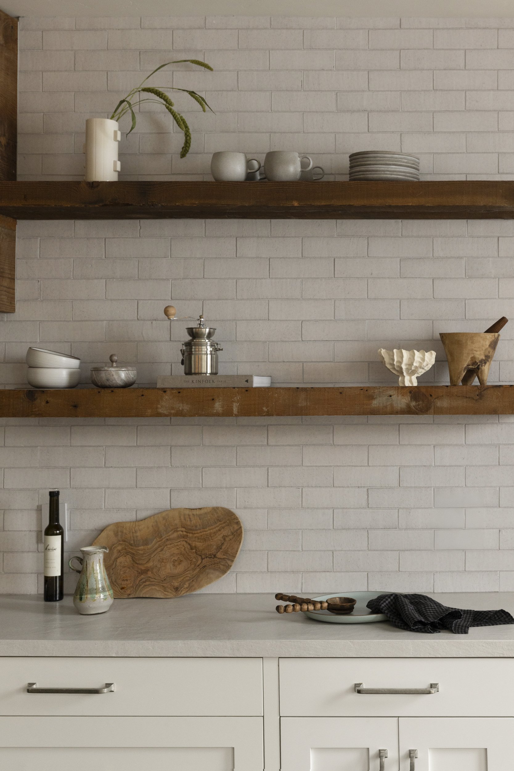 White kitchen counter with decorative items including a large wooden tray, a ceramic pitcher, a bottle of wine, and a plate. White cabinets with silver handles below. Two wooden shelves above with stacked plates, cups, a vase with greenery, and other ceramic and glass items against a white brick wall.