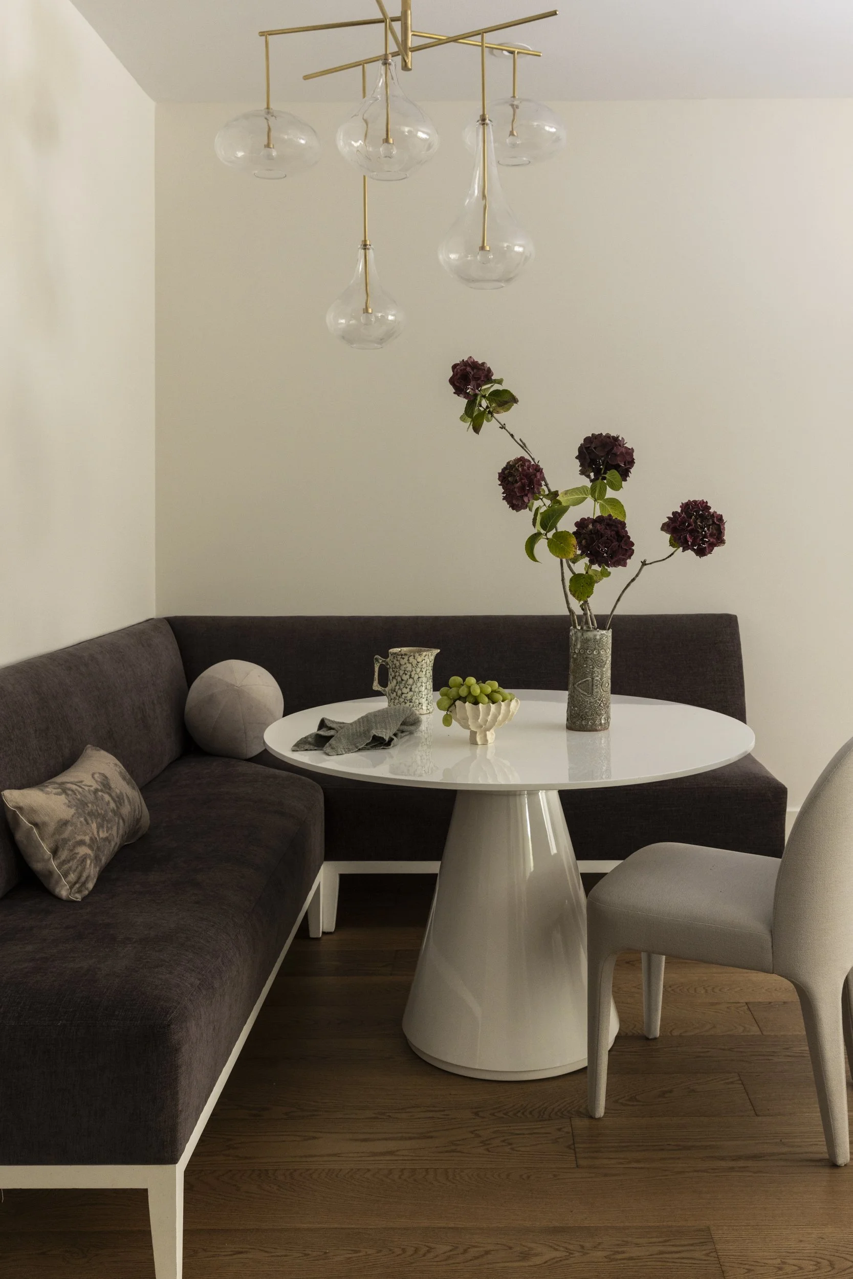 A round white dining table with a modern vase of pink and purple flowers, a small bowl of green grapes, a decorative cup, and napkins. Surrounding the table are a gray upholstered chair and a brown sectional sofa with patterned pillows. Above, there 
