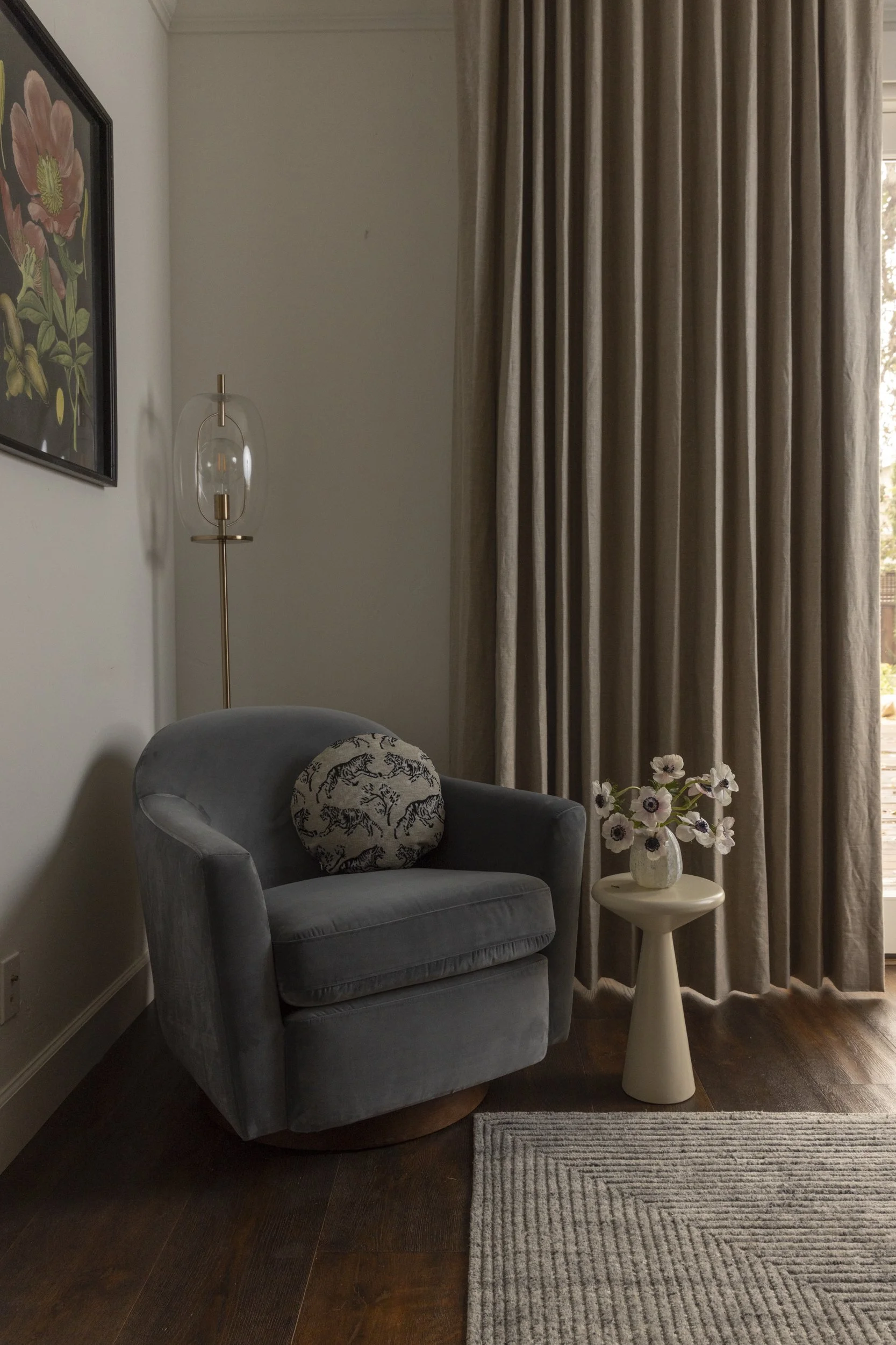 A cozy living room corner featuring a blue velvet armchair with a round pillow decorated with a black and white tiger pattern. Next to the chair is a small white side table holding a white vase with pink and white flowers. Behind the chair, beige cur