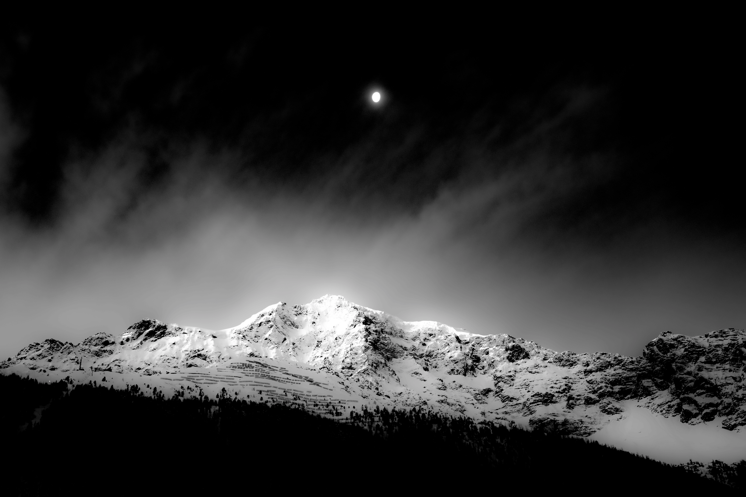 Black and white photograph of a snow-covered mountain with a bright moon in the dark sky above.