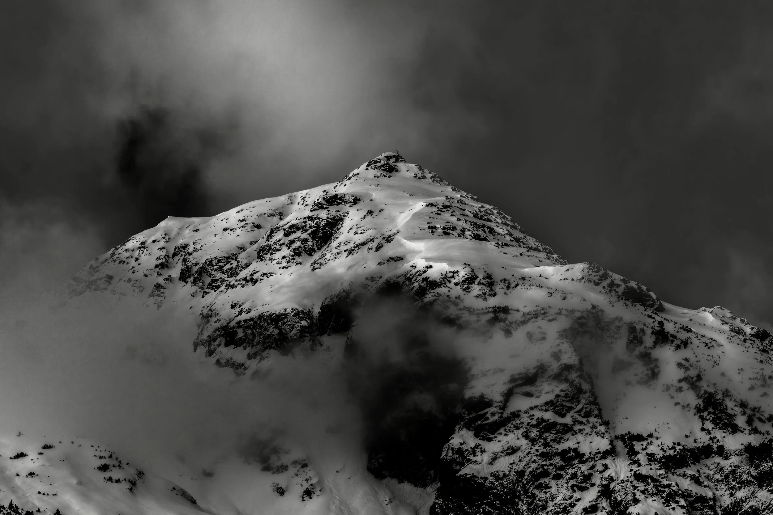 Snow-covered mountain peak with a cross at the summit, dark cloudy sky, and rugged slopes, in black and white.