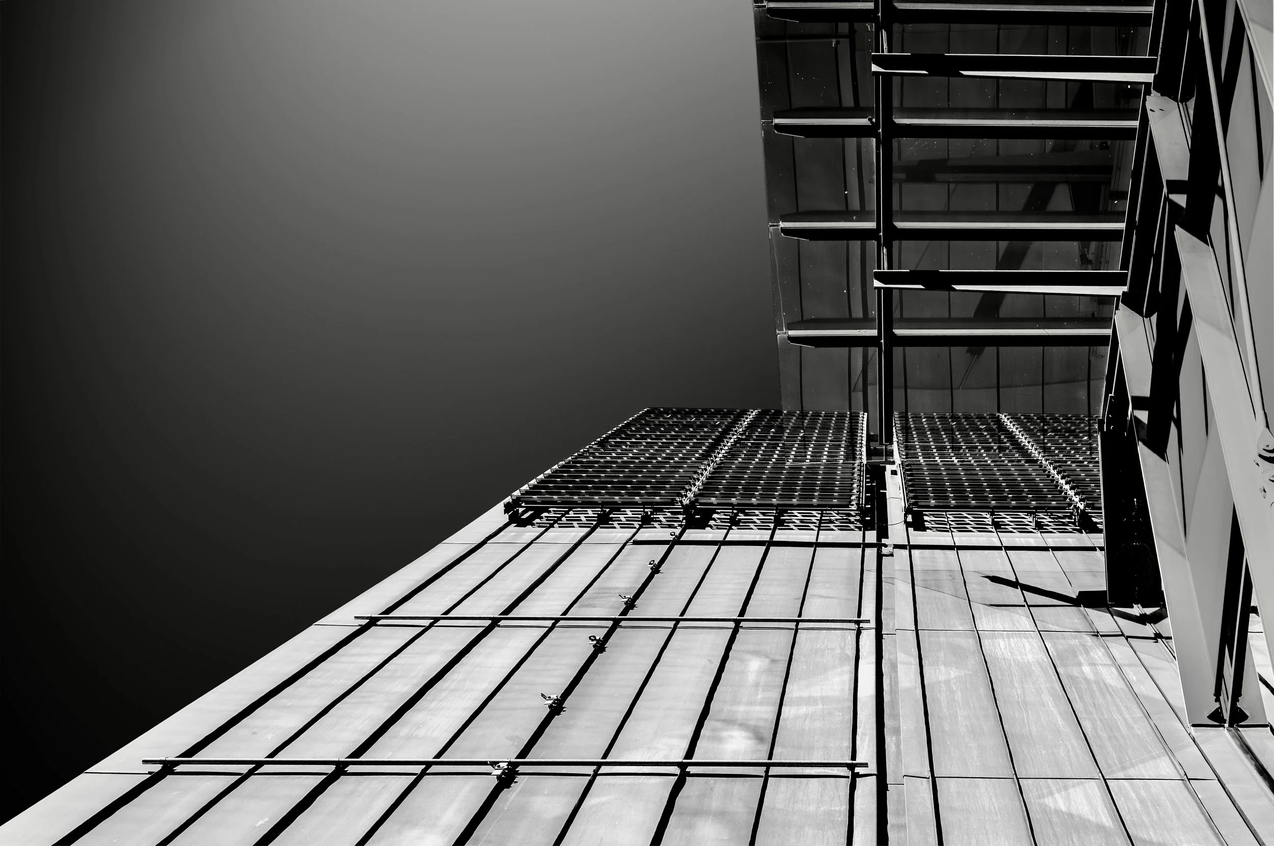 A black and white photo of a modern building with a glass facade, viewed from below. The building has a grid-like metal framework and multiple horizontal and vertical elements, with a clear sky in the background.