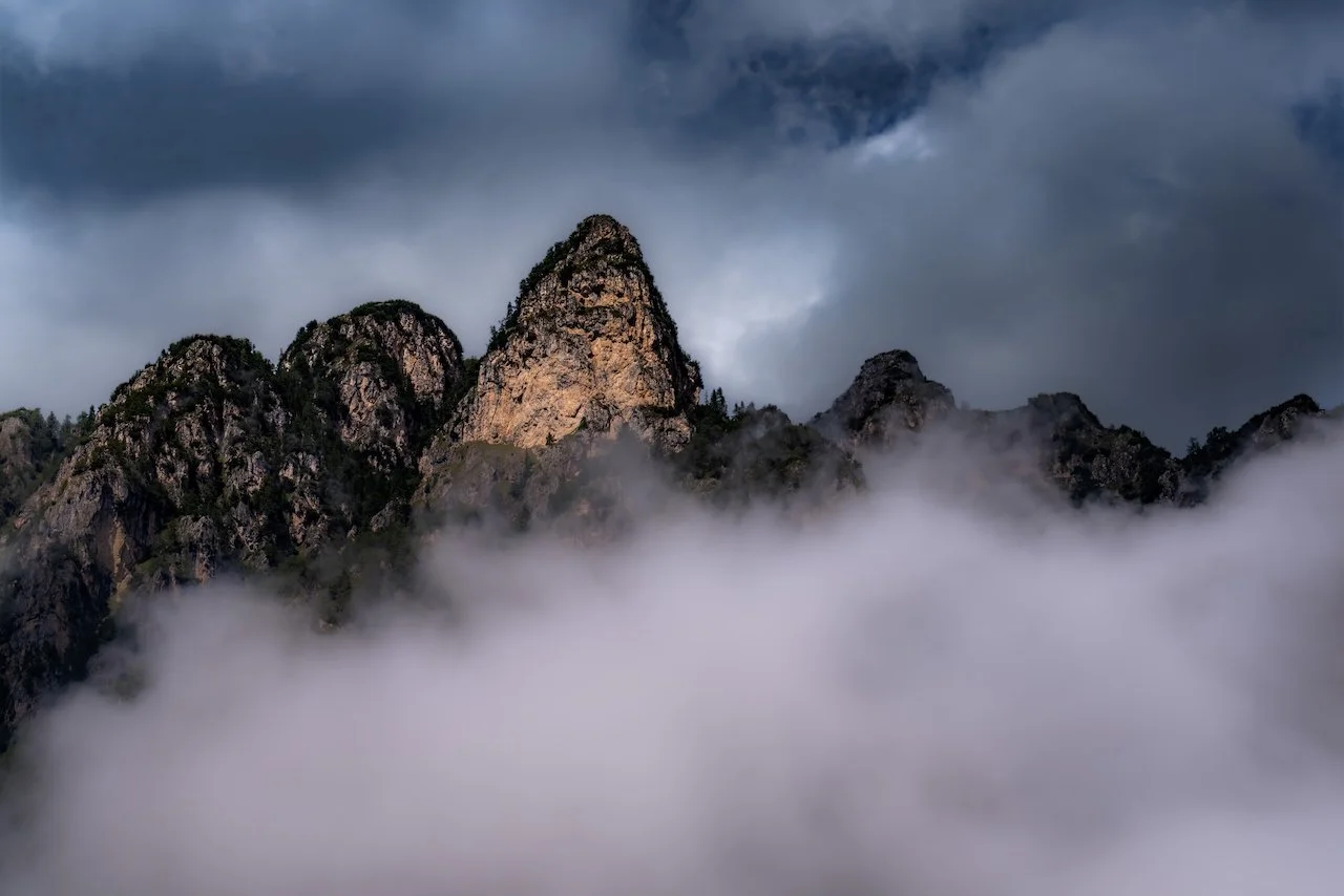 Mountain peaks emerging from clouds with dark, cloudy sky overhead.