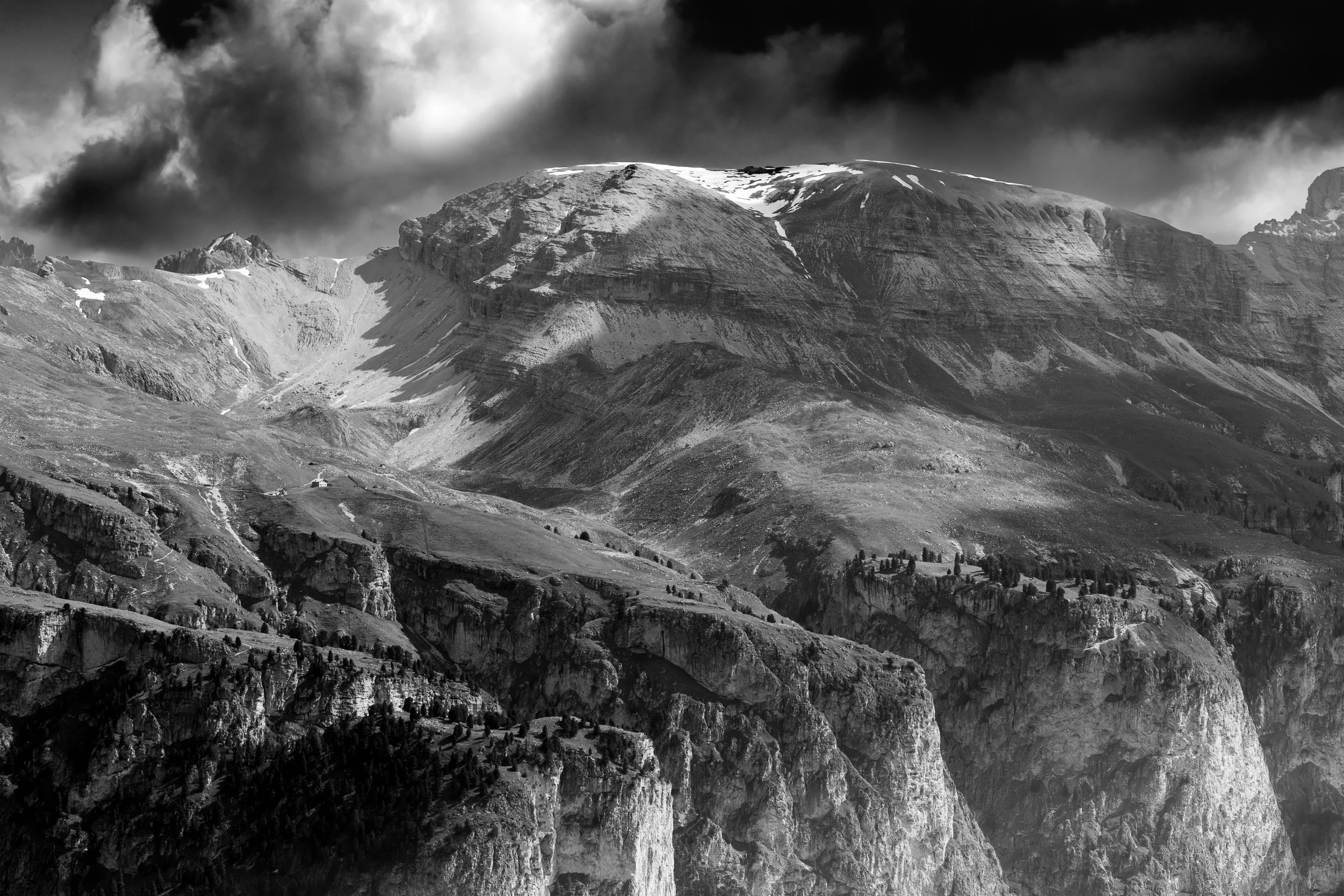 Black and white photograph of rugged mountain landscape with steep cliffs, snow patches, and clouds overhead.
