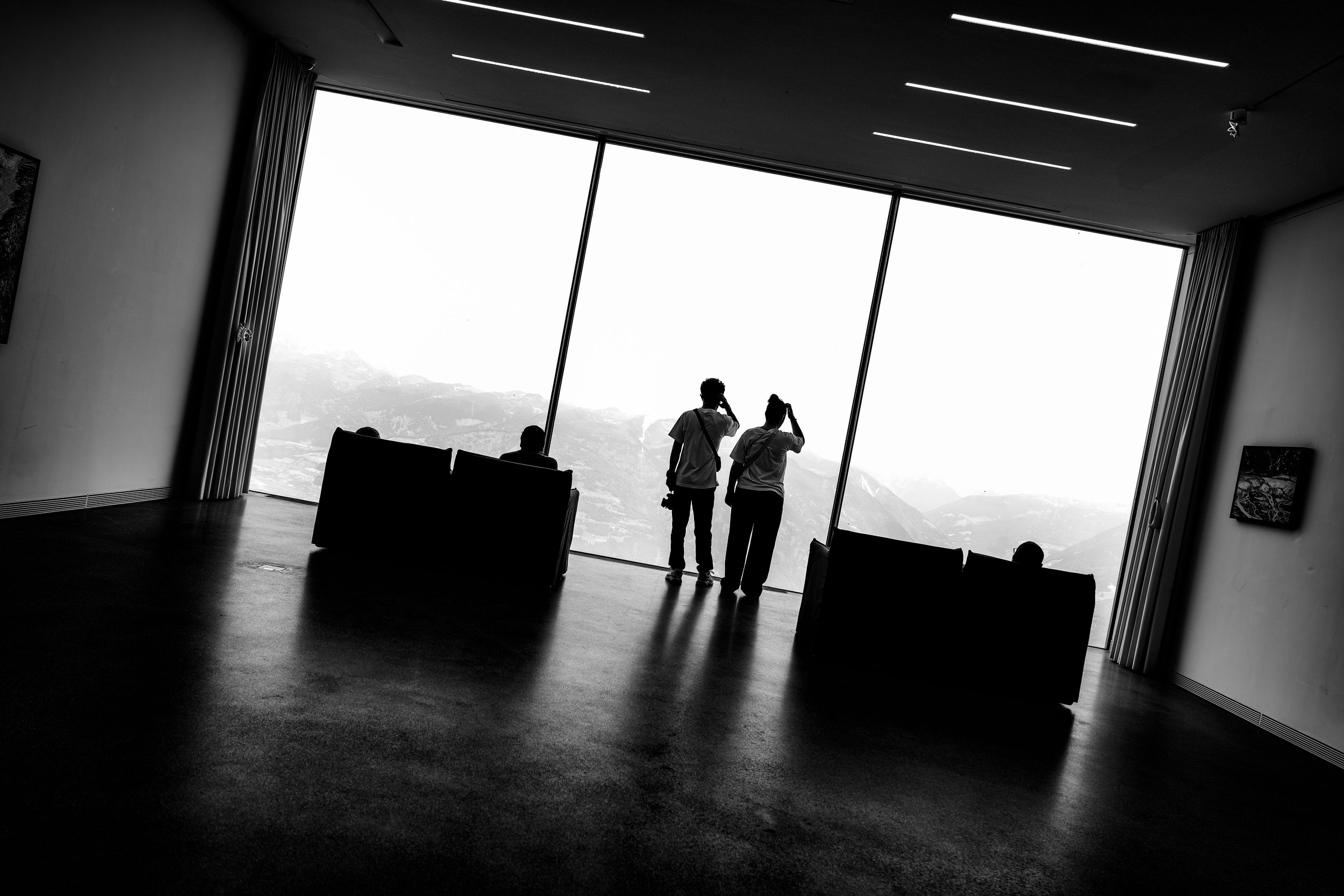 Silhouettes of three people standing in front of large windows with mountains visible outside in a modern indoor space.