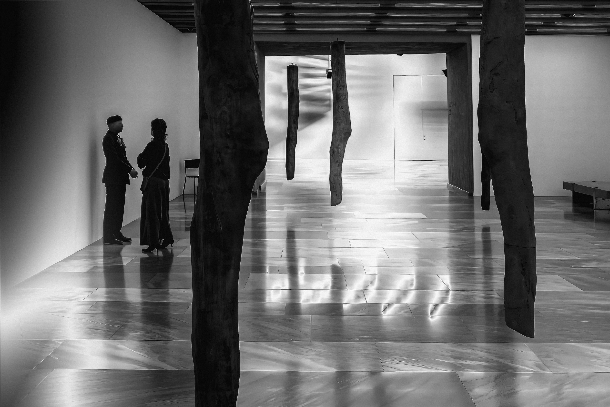 Black and white photo of two people, a man in a suit and a woman with a bag, standing and conversing in a modern, minimalist interior with wooden flooring and vertical wooden sculptures.