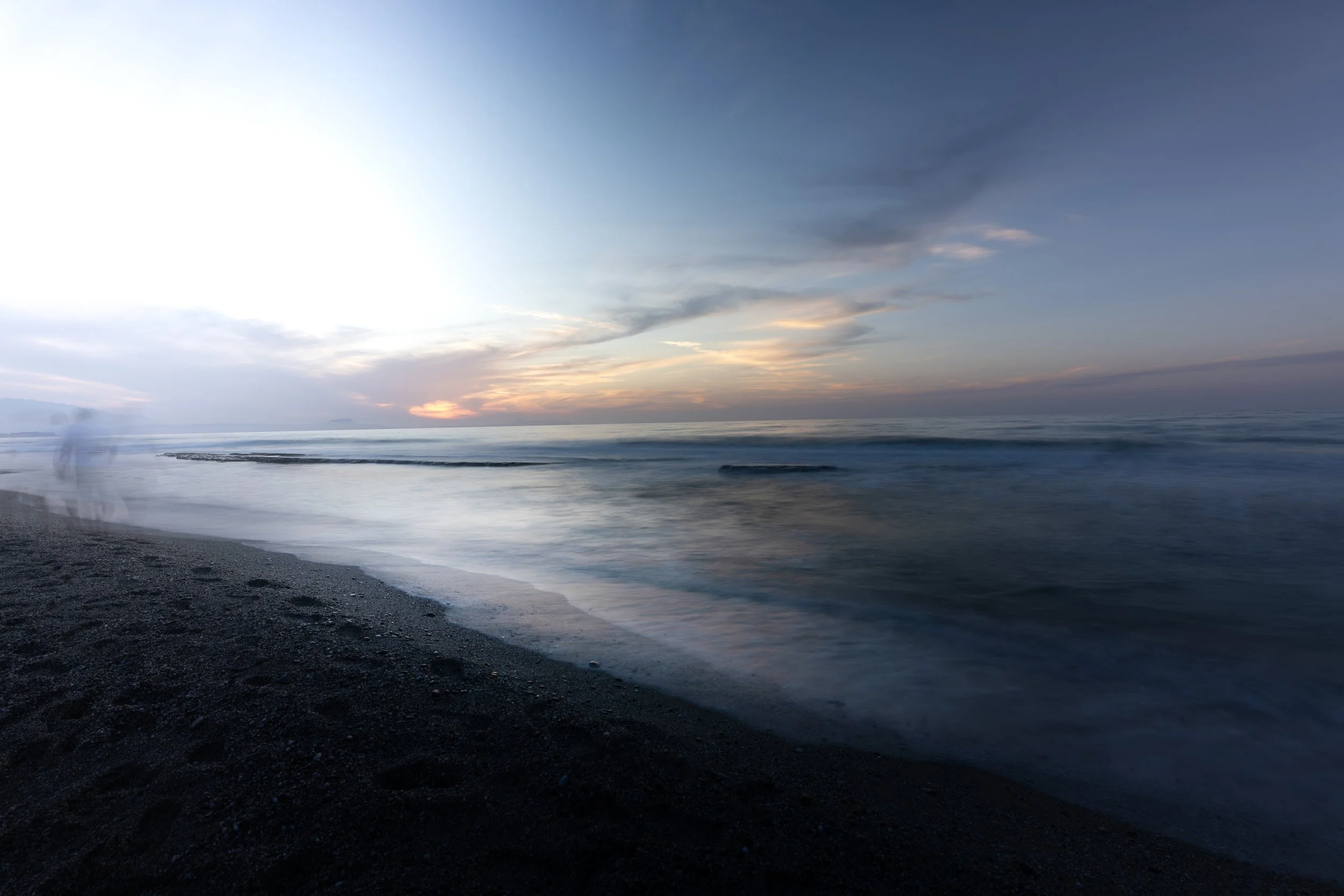 A peaceful beach at sunset with dark sand, calm ocean waves, and a sky with clouds displaying soft pastel colors.