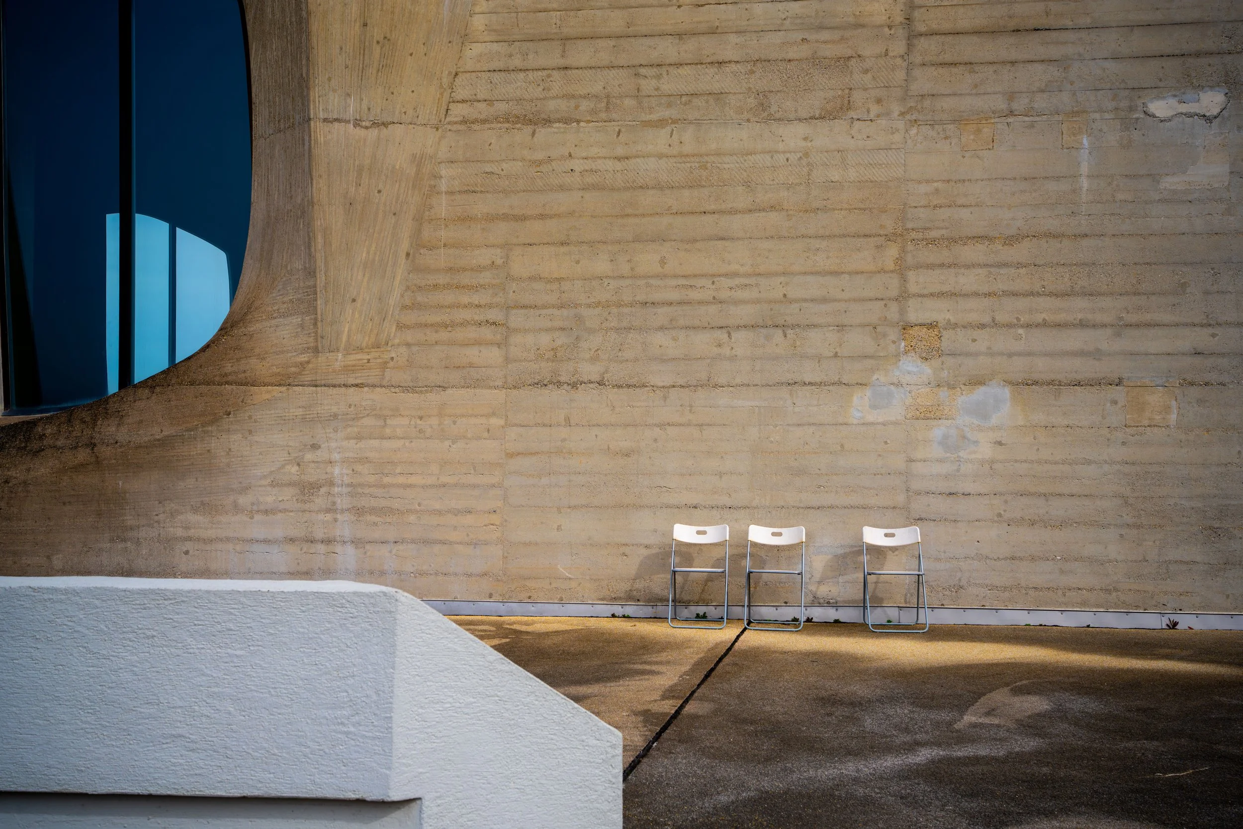 Three white chairs placed side by side against a large, textured concrete wall with curved architectural features and a window on the left side.