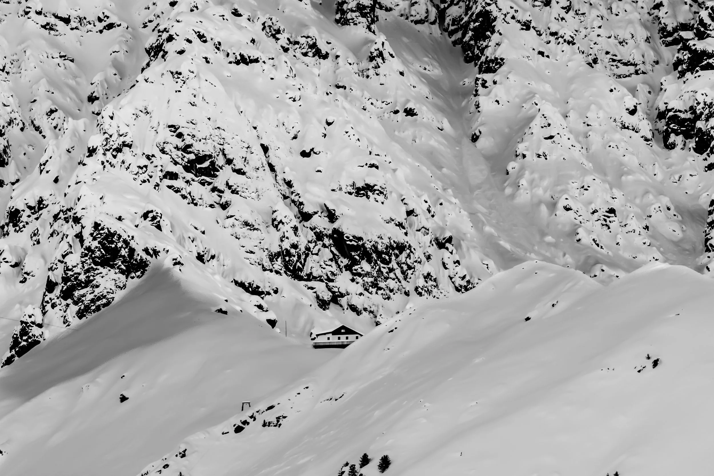 A snow-covered mountain with a small house beneath the slopes and a few trees in the foreground.