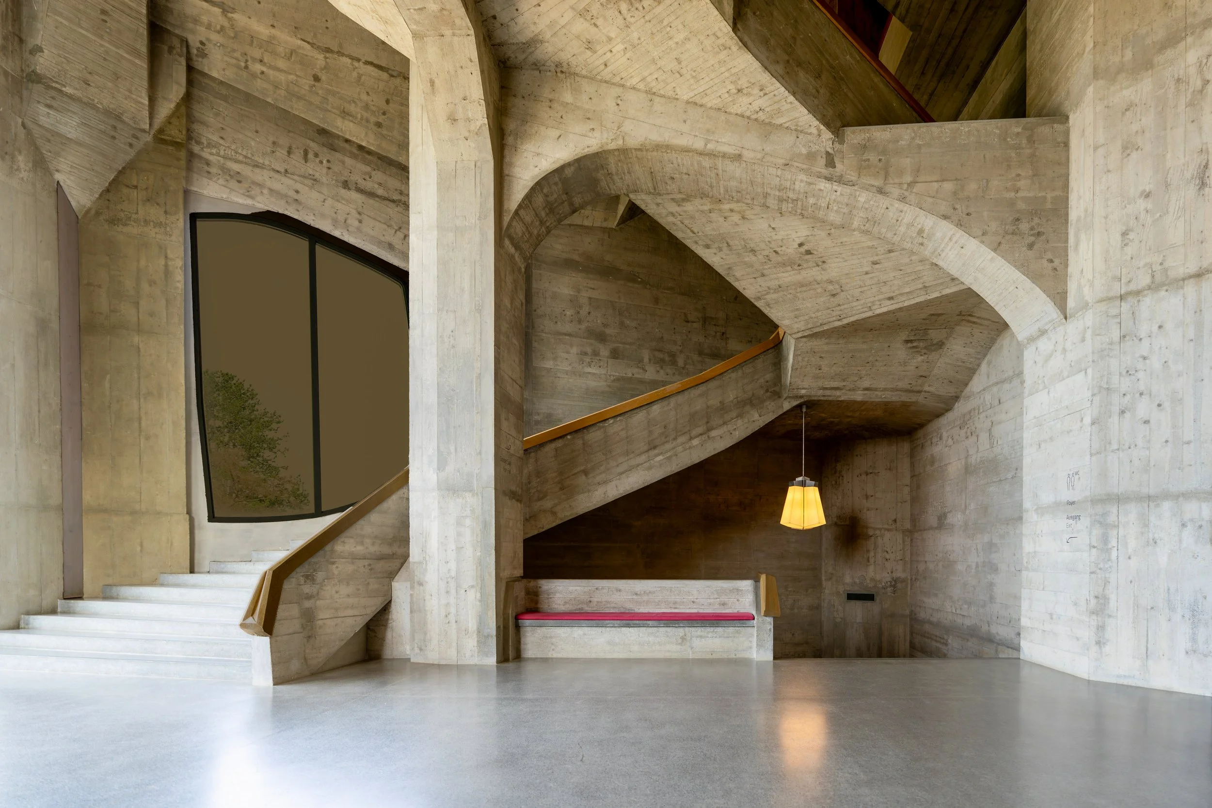 Interior view of a modern building with curved concrete walls, a staircase with wooden handrails, large curved window, hanging light, and text on wall.
