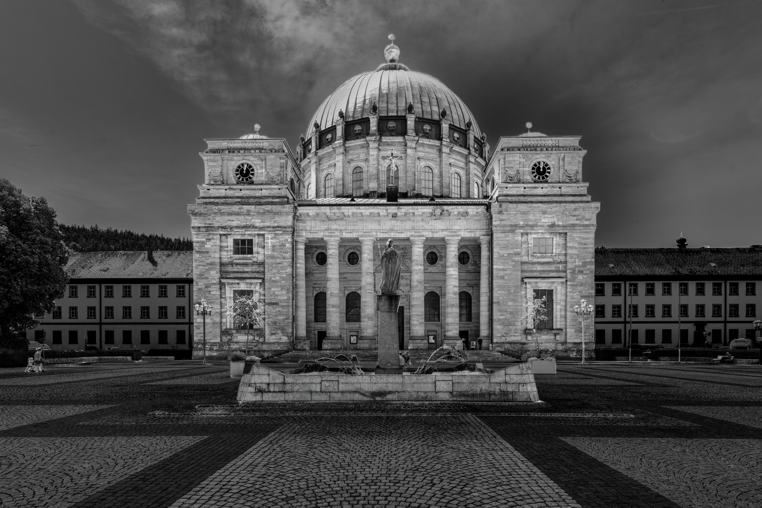 Black and white photograph of a historic building with a large dome, clock towers, and a statue in front, surrounded by a paved square and some trees.