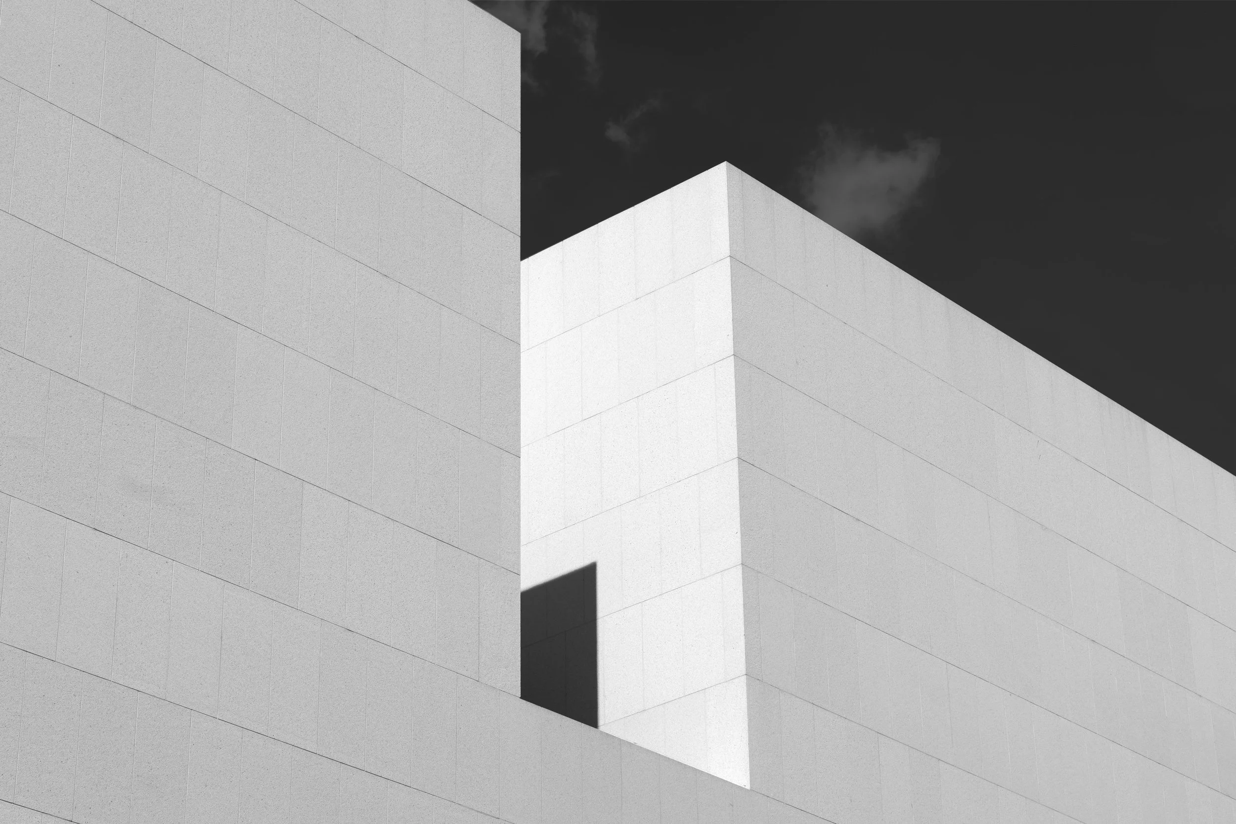 Black and white photo of a modern building with sharp angles and flat surfaces, set against a dark sky with some clouds.