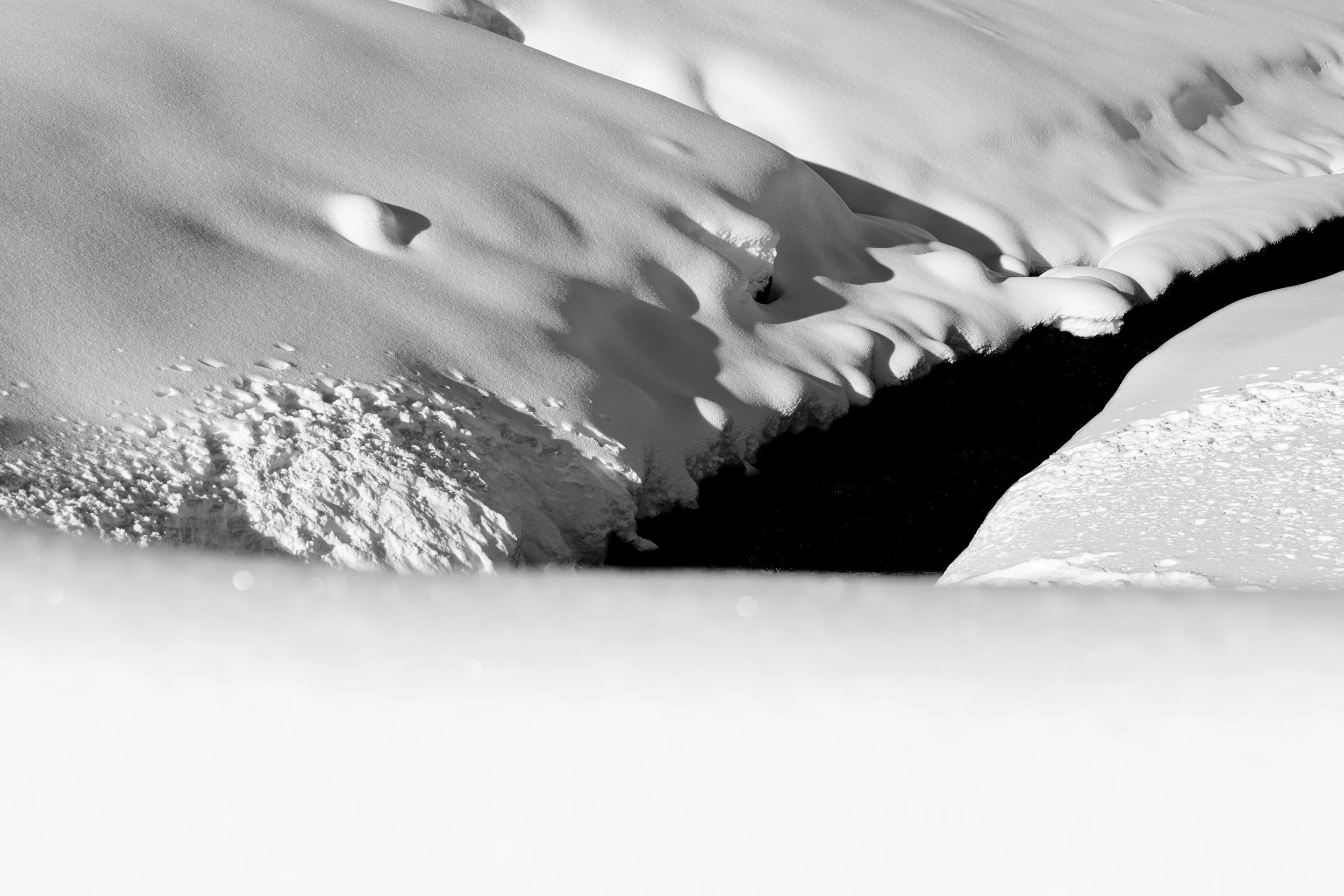 Black and white photo of snow-covered landscape with smooth snowdrifts and a dark stream or river running through the snow.