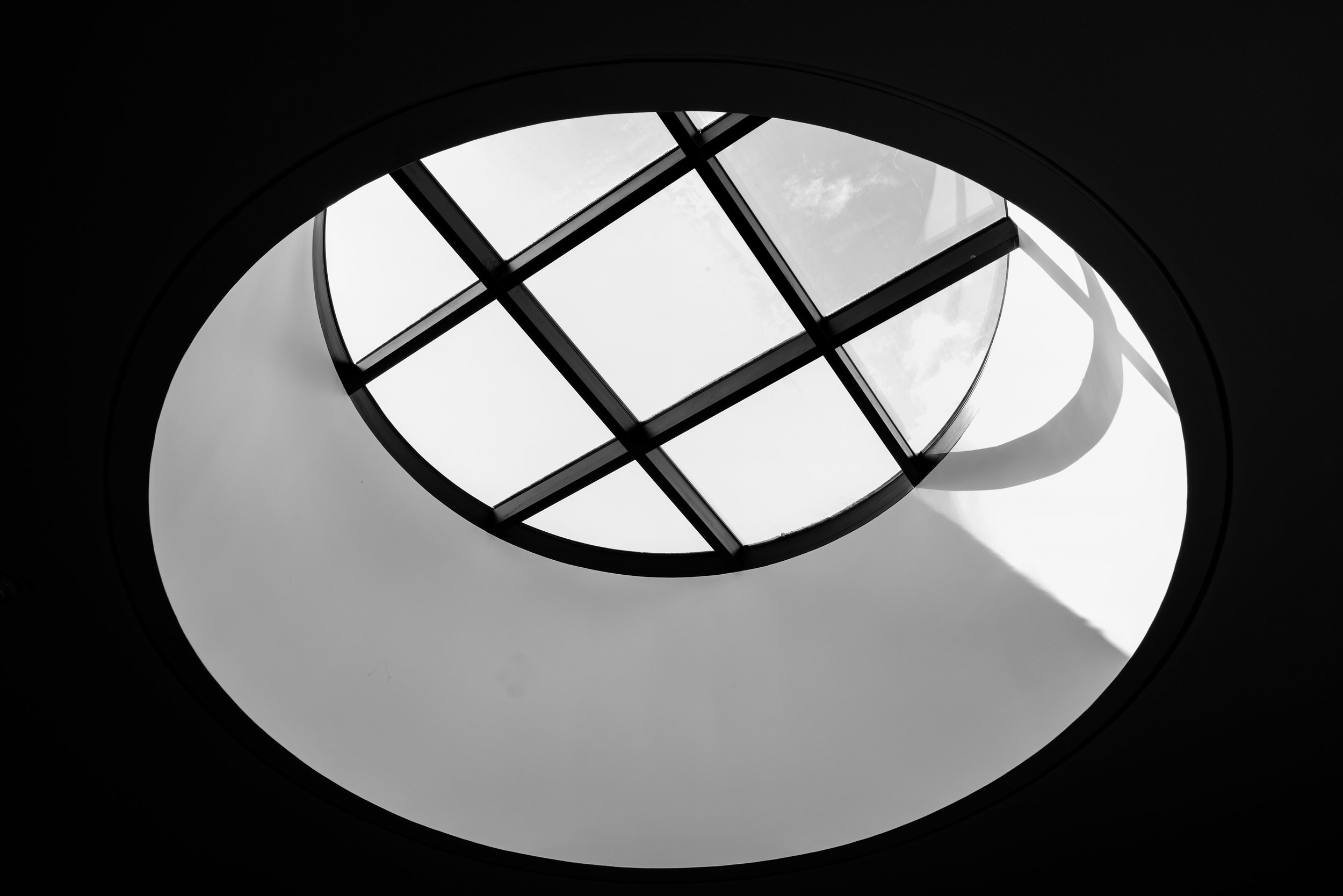 Black and white photo taken from below of a glass window in a circular opening, with the sky and some clouds visible through the window.