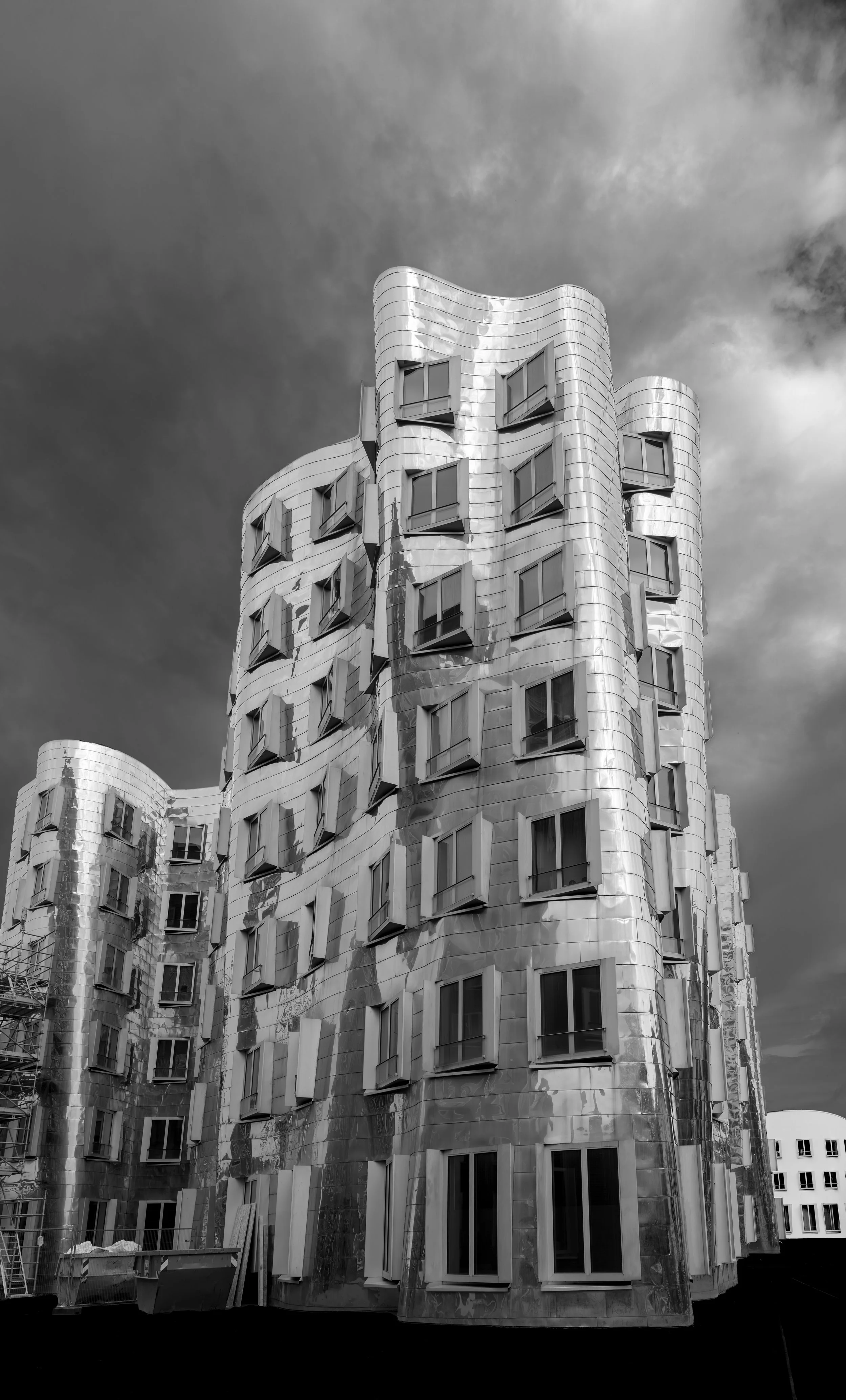 An undulating modern building with a reflective metal surface and irregularly arranged windows, set against a cloudy sky.