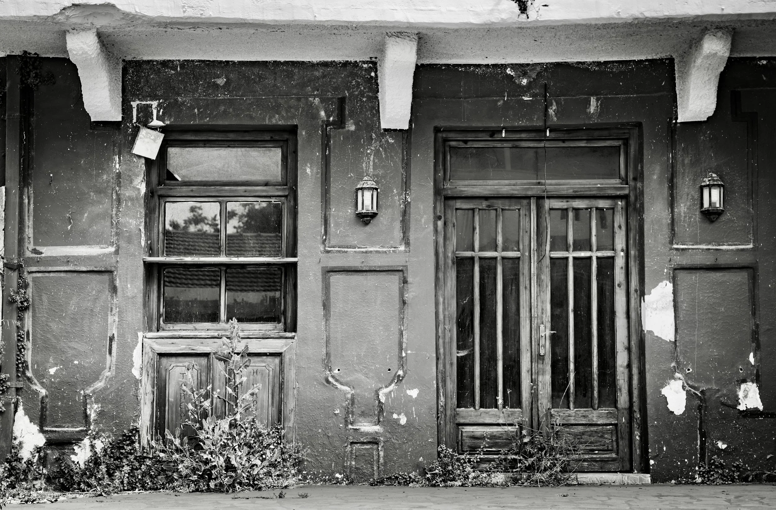 Black and white photo of an old, worn building facade with two windows and a door. The windows have wooden frames and are partially covered in dirt. The door is wooden with panels. There are two wall-mounted lanterns on either side of the door, and t