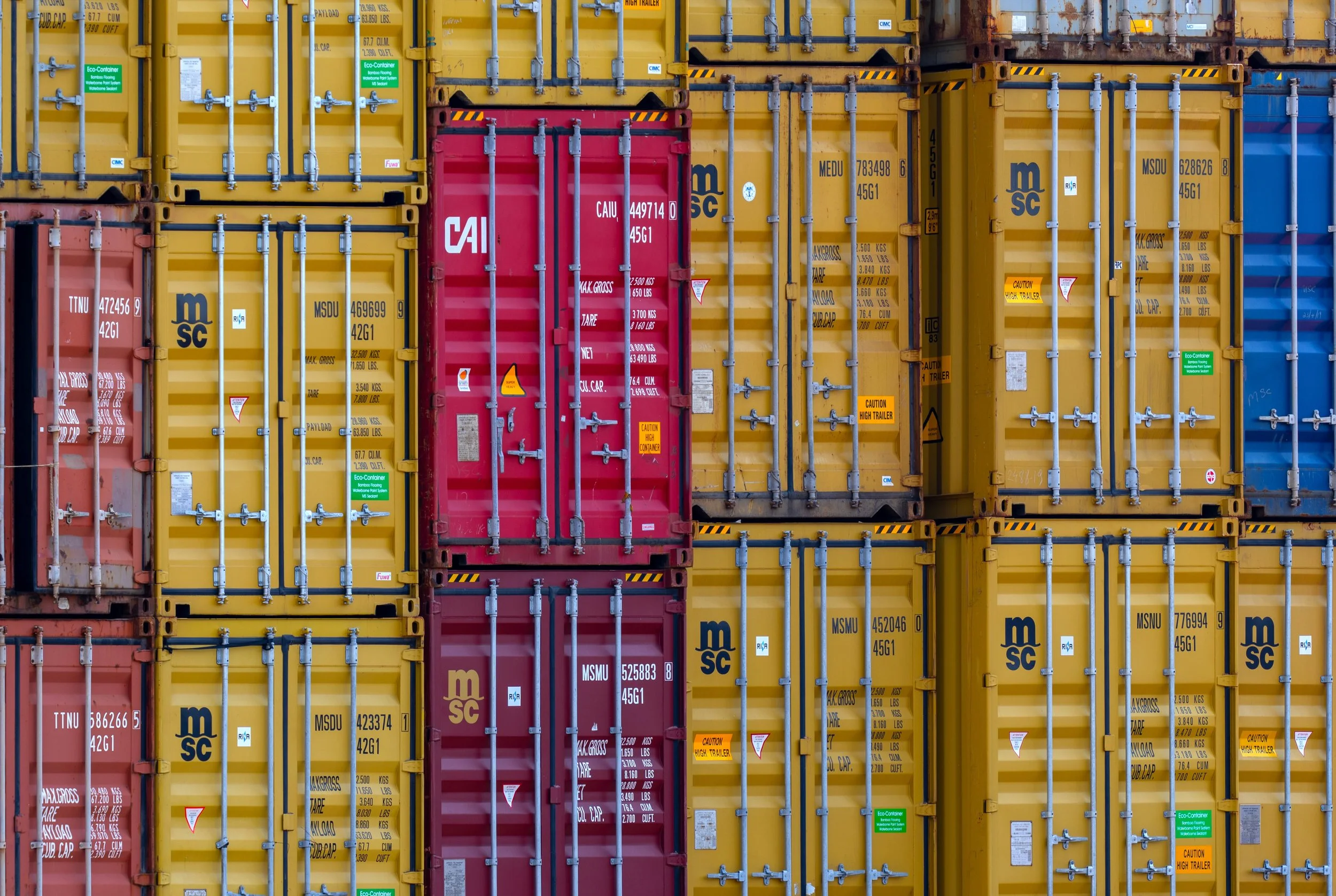 A close-up view of stacked colorful shipping containers, primarily yellow, with some red and blue, secured with metal locking bars at a port or storage yard.