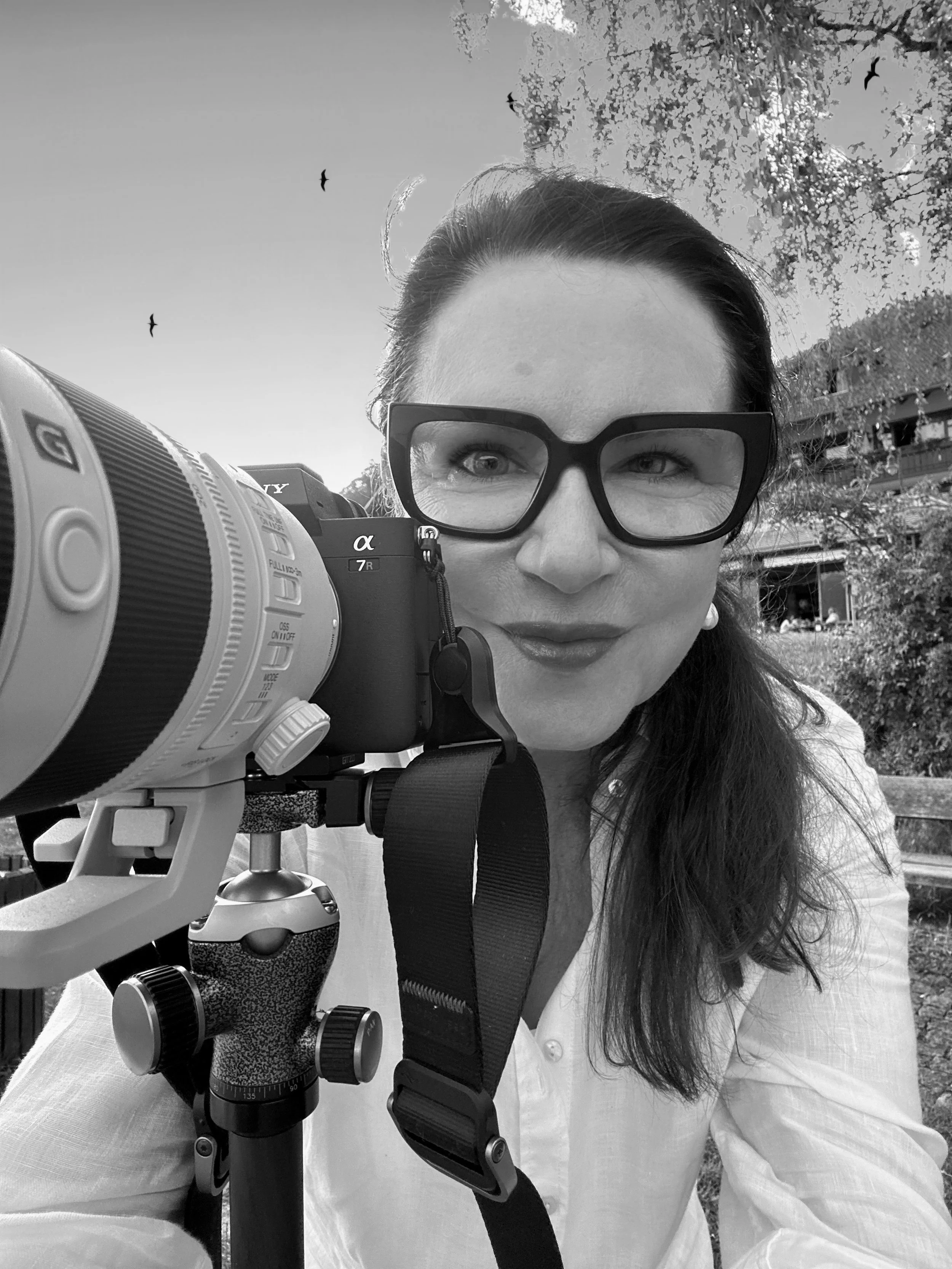 Black-and-white photo of a woman with glasses taking a photo with a camera on a tripod outdoors, with trees and buildings in the background, and birds flying in the sky.