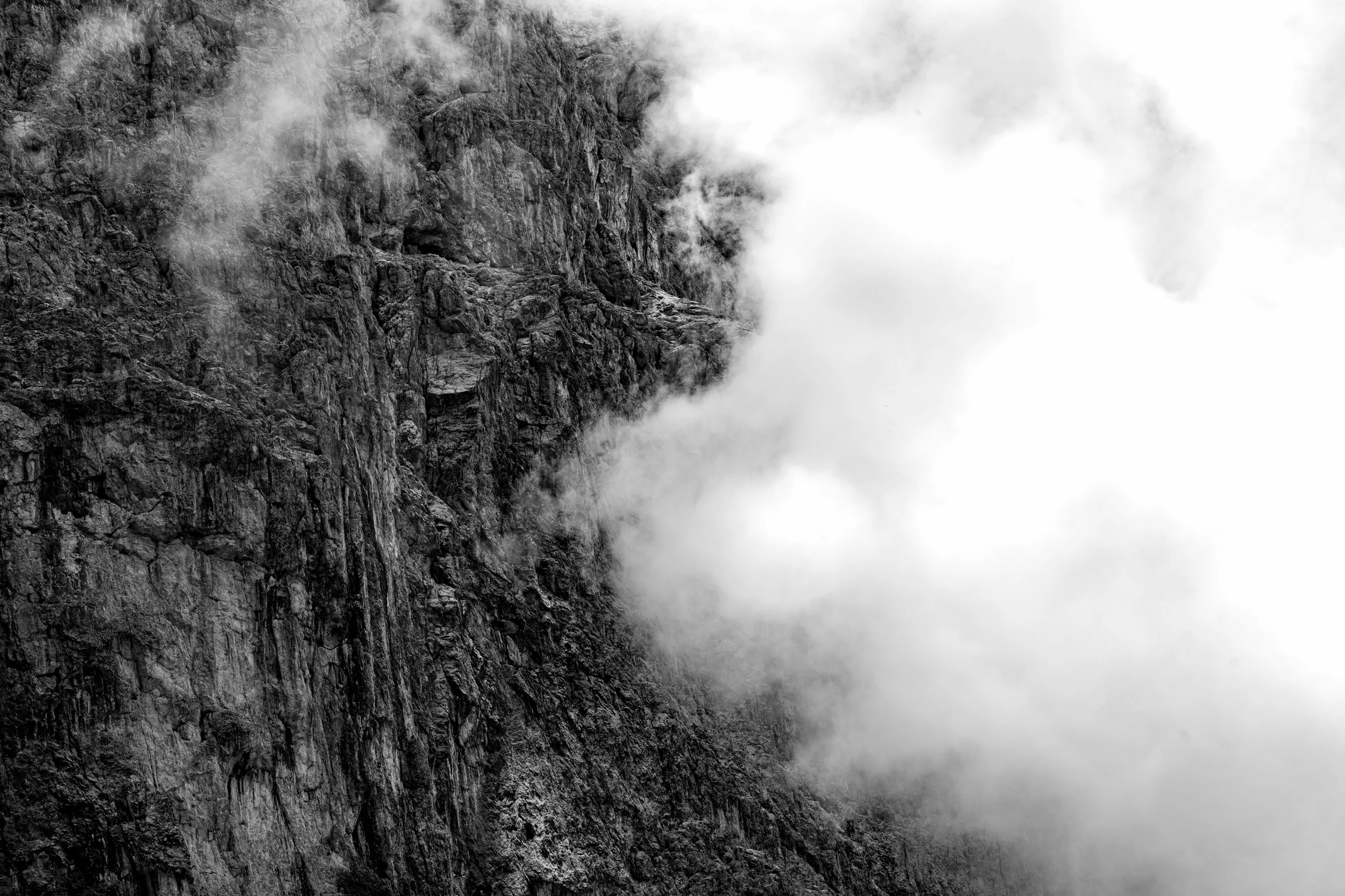 Black and white photo of a rocky mountain face with clouds or mist obscuring parts of it.