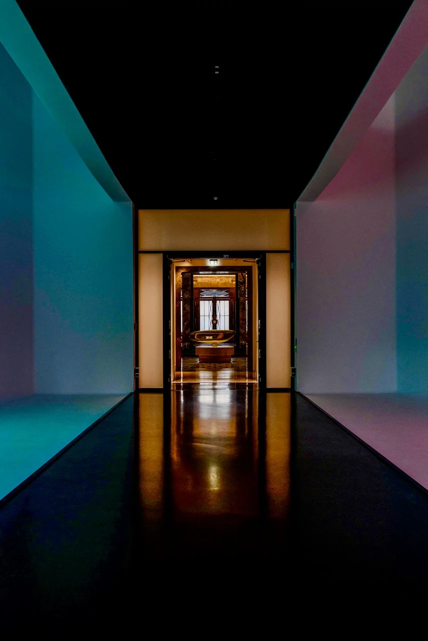 A dimly lit hallway leading to an ornate door, reflecting warm light, with colorful wall lighting on each side.