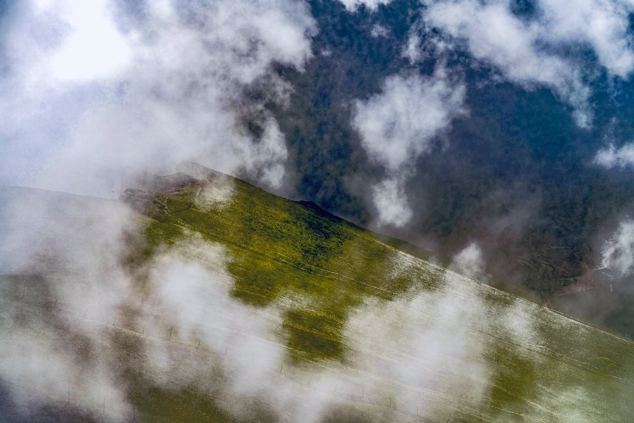 Mountain with patches of green grass and misty clouds surrounding it.