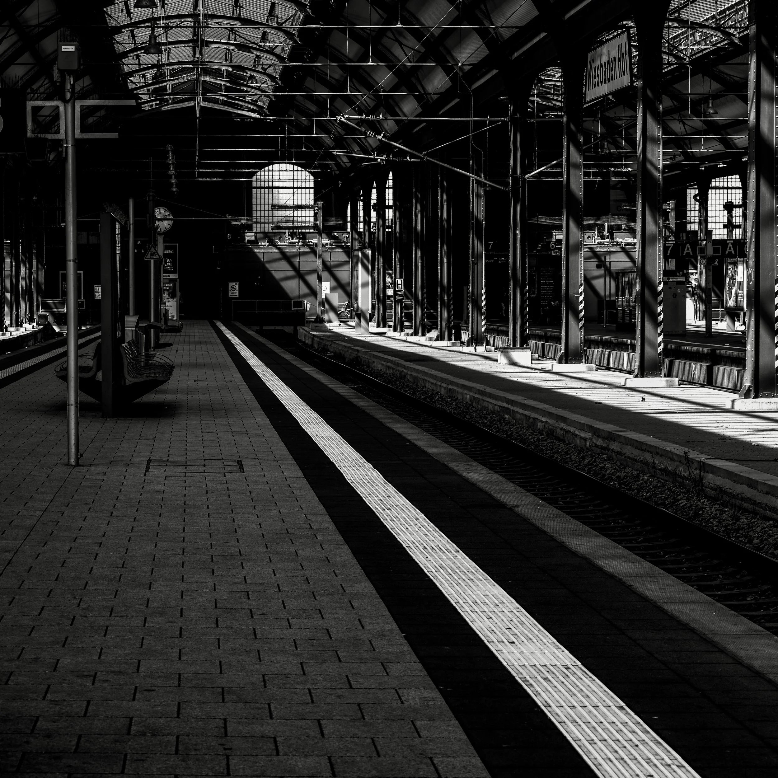 Black and white photo of a train station platform with benches, tracks, and a sign that reads 'Mesadaet' under a large arched glass roof, with shadows cast on the ground.
