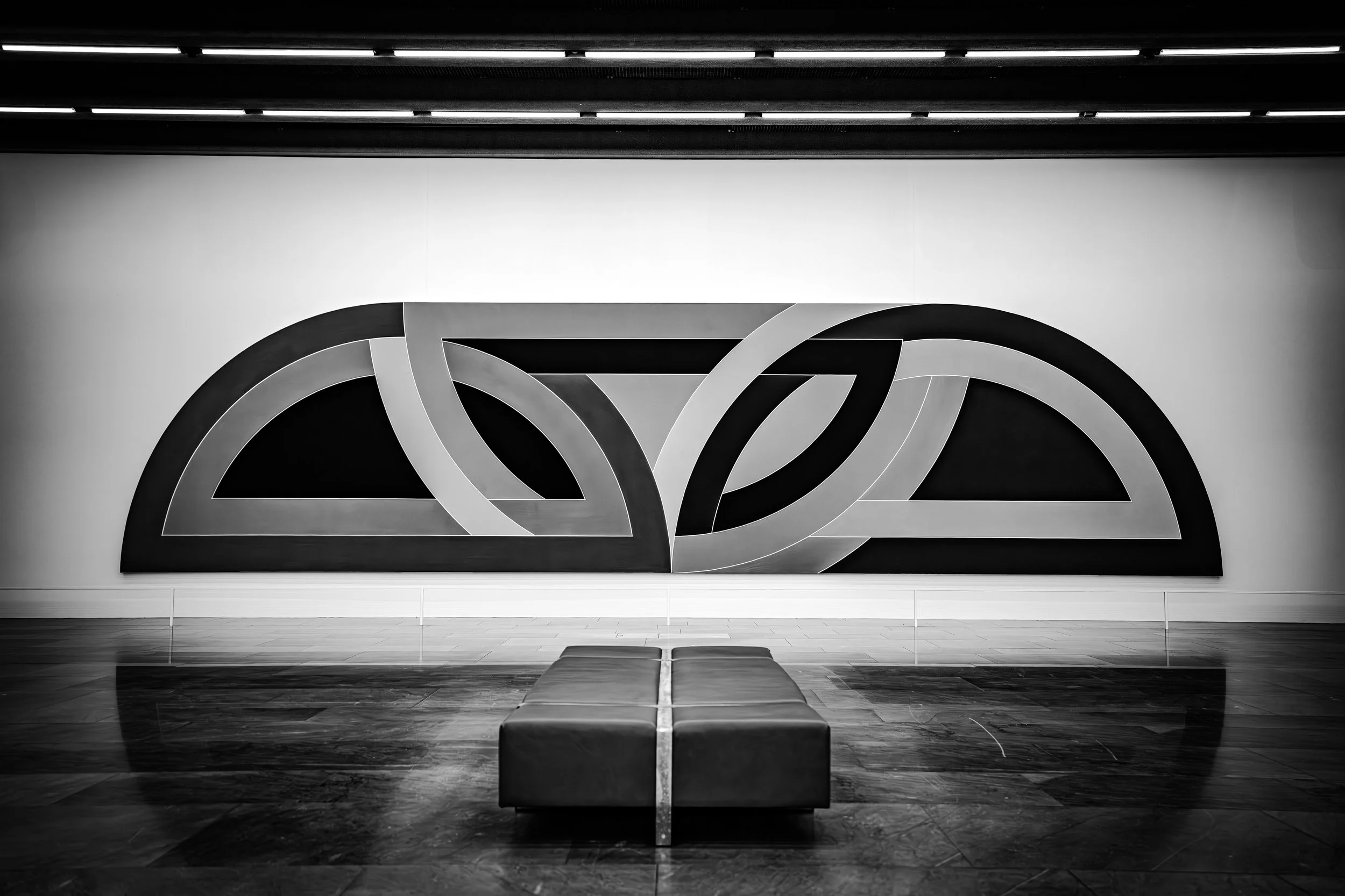 Black and white abstract artwork on a wall with curved and angular shapes, a bench in the foreground, and ceiling lights overhead.