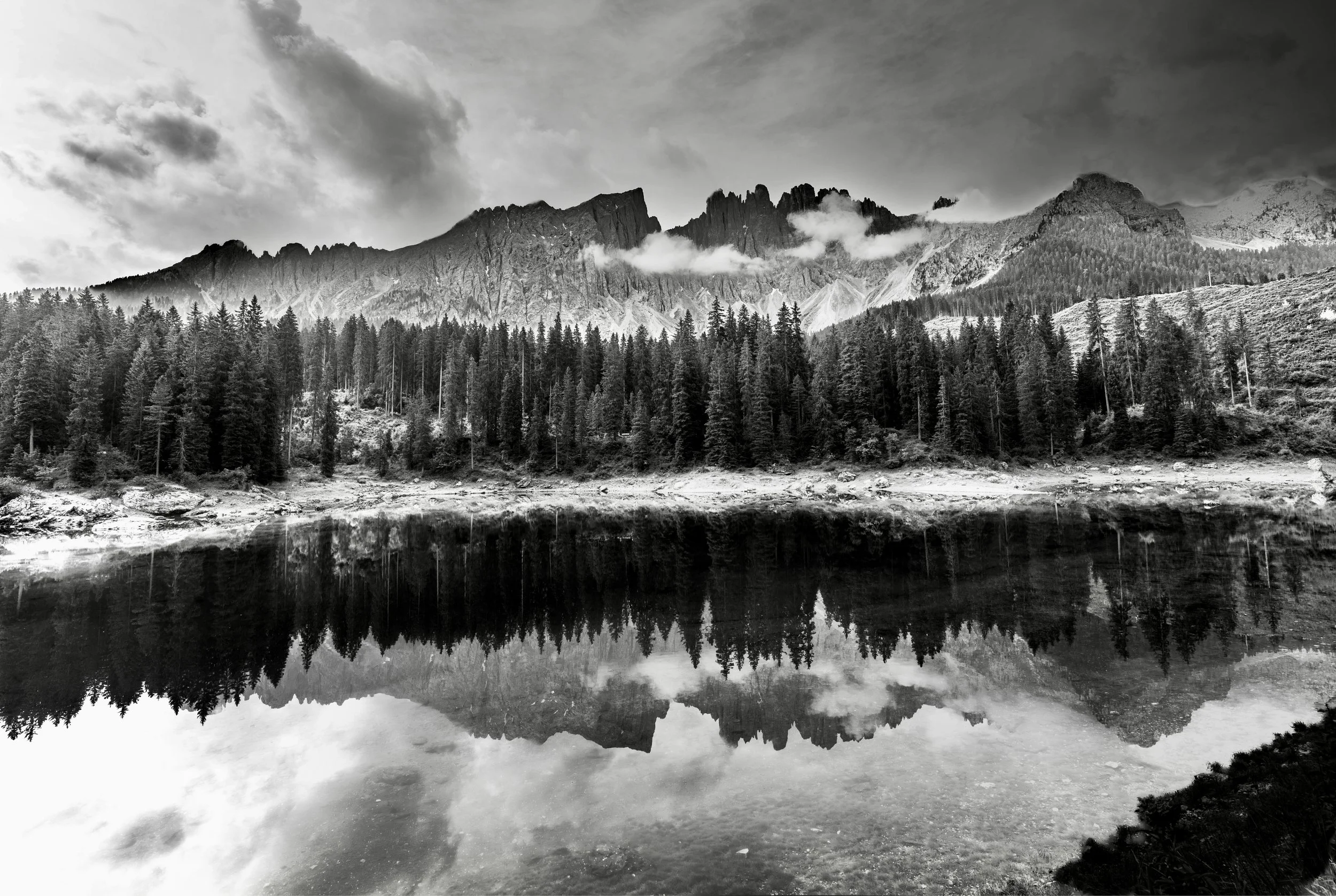 Black and white photo of a mountain range with clouds around its peaks, a forest in the foreground, and a reflection of the mountains and forest in a body of water.