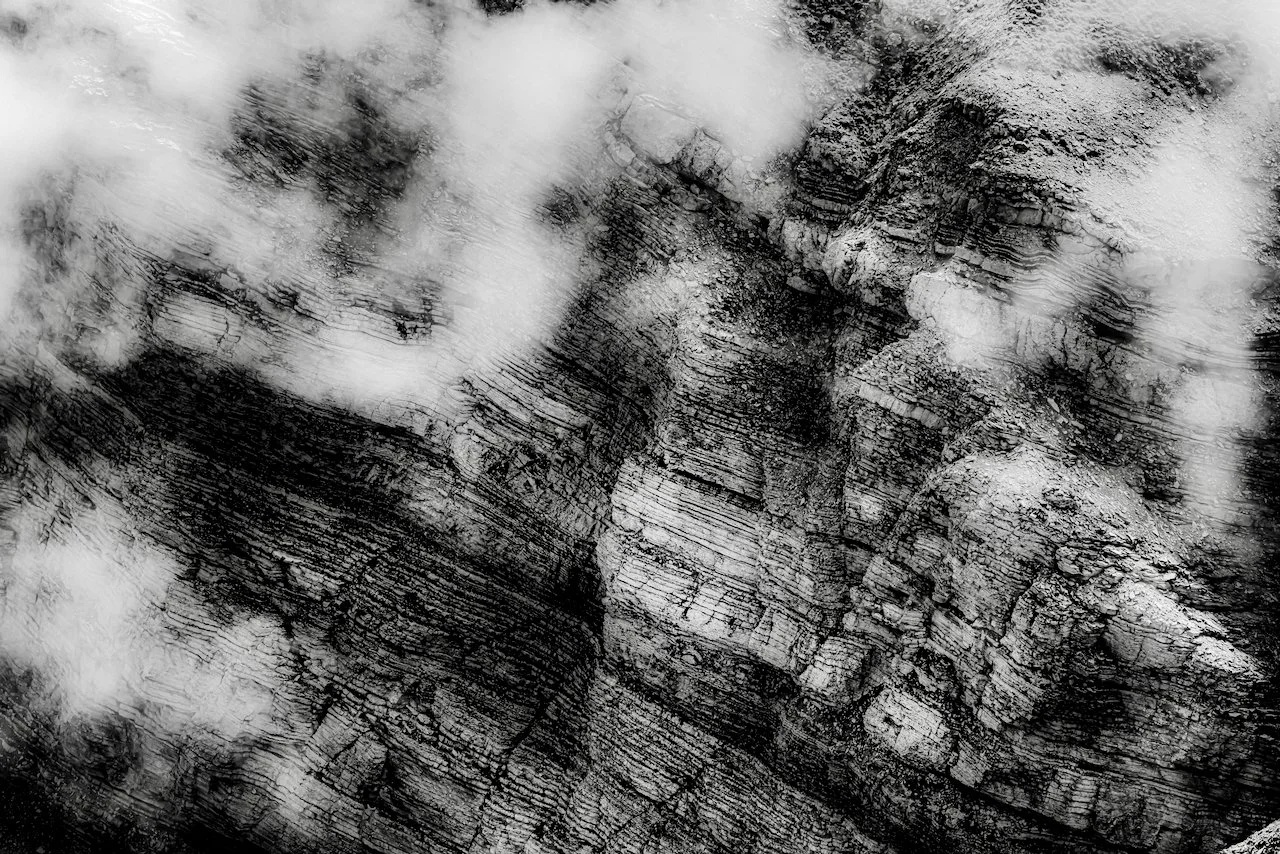 Black and white aerial photograph of a cloud-covered, rugged mountain landscape with layered rock formations.