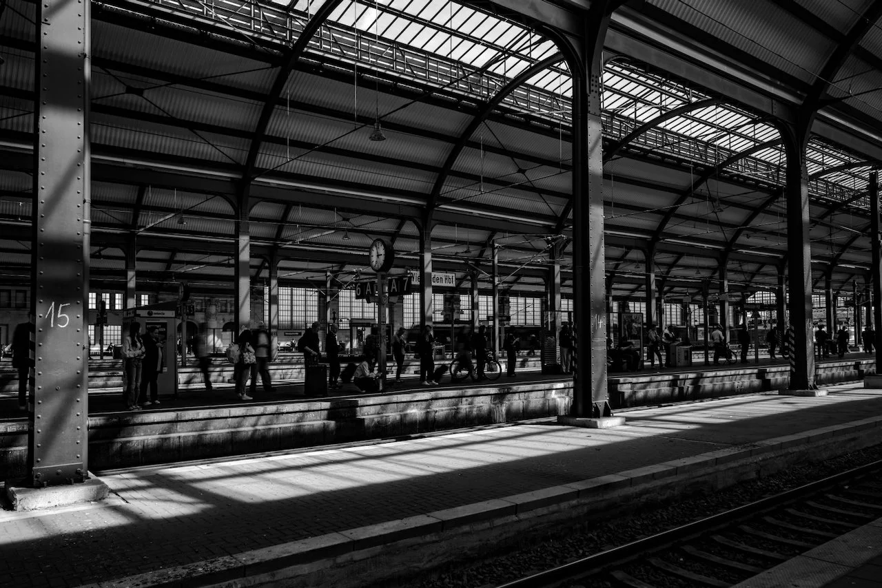 Black and white photo of a train station with waiting passengers on the platform, metal roof, and train tracks in the foreground.