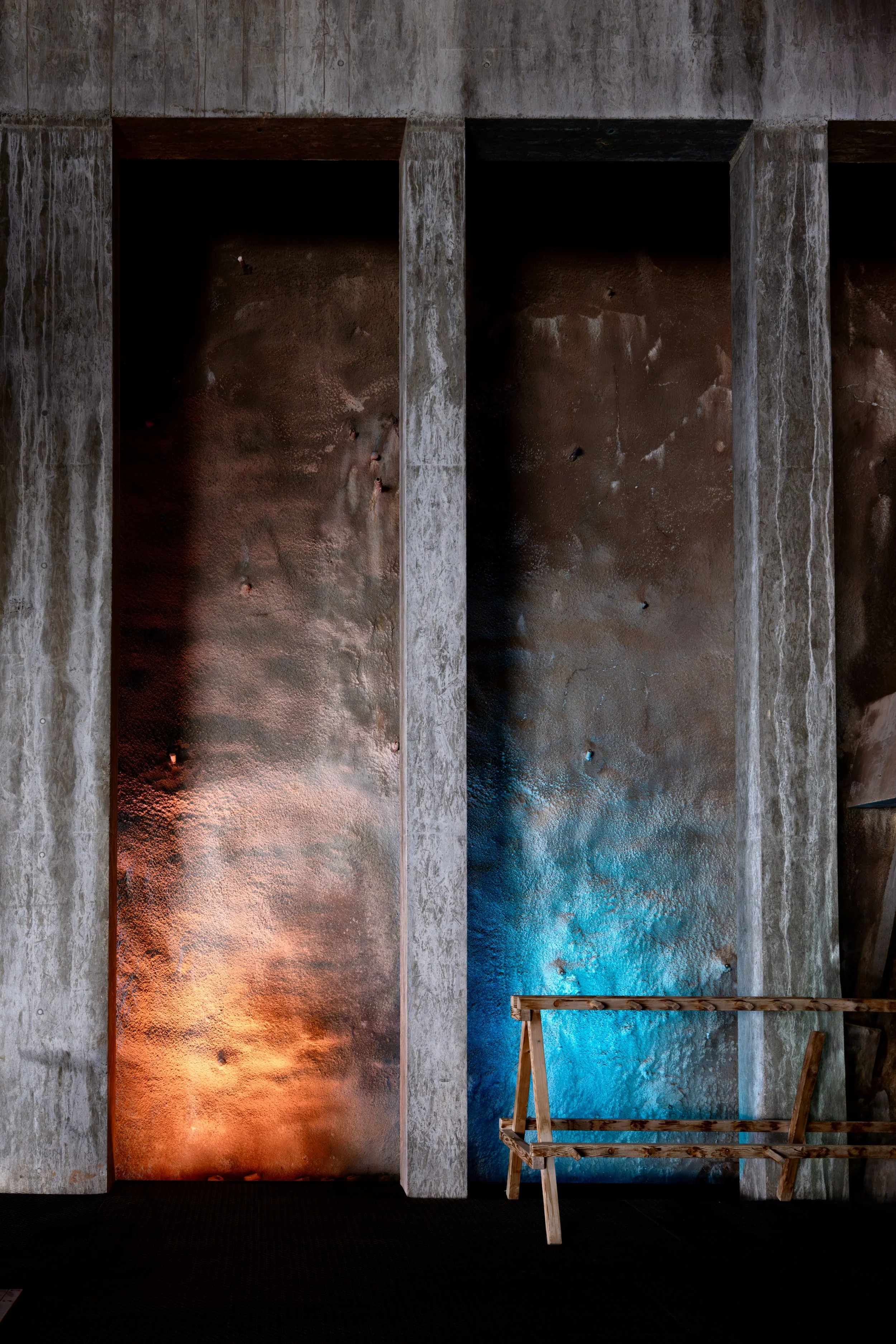 Concrete wall with three vertical backlit panels in orange, blue, and rusty colors; wooden stair bench in the foreground.