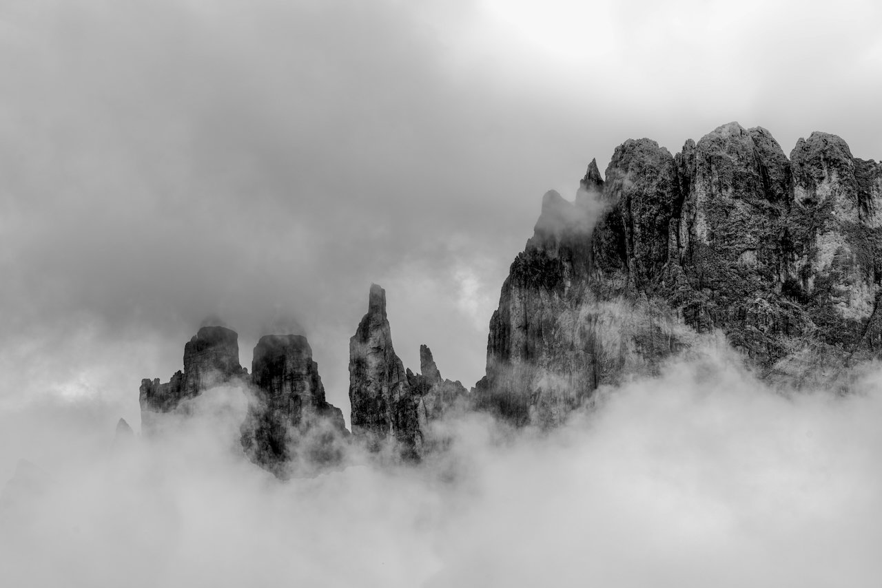 Black and white photograph of tall rugged mountain peaks surrounded by clouds or fog.