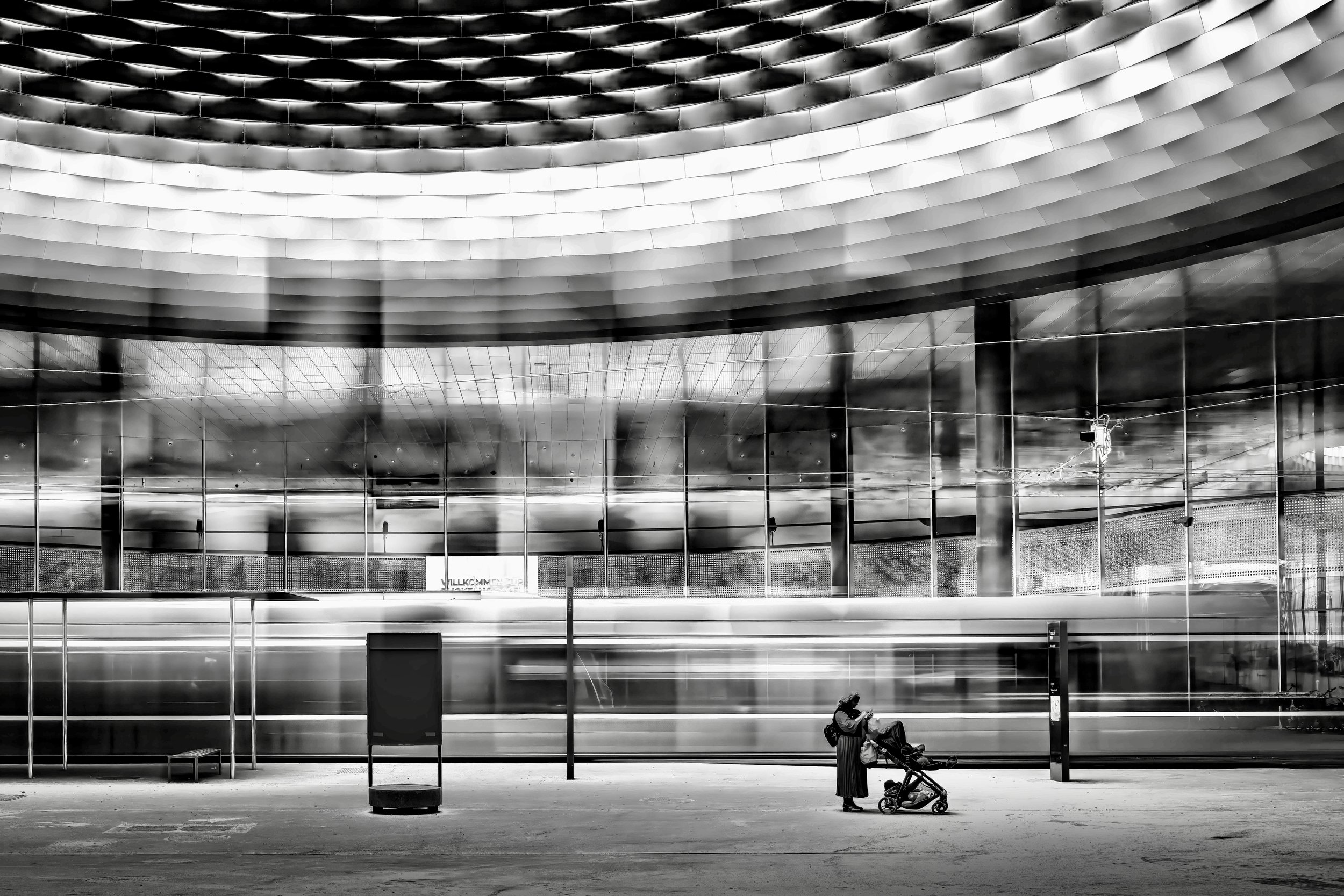 A woman with a stroller waiting at an airport terminal, with modern architecture and glass walls in the background.