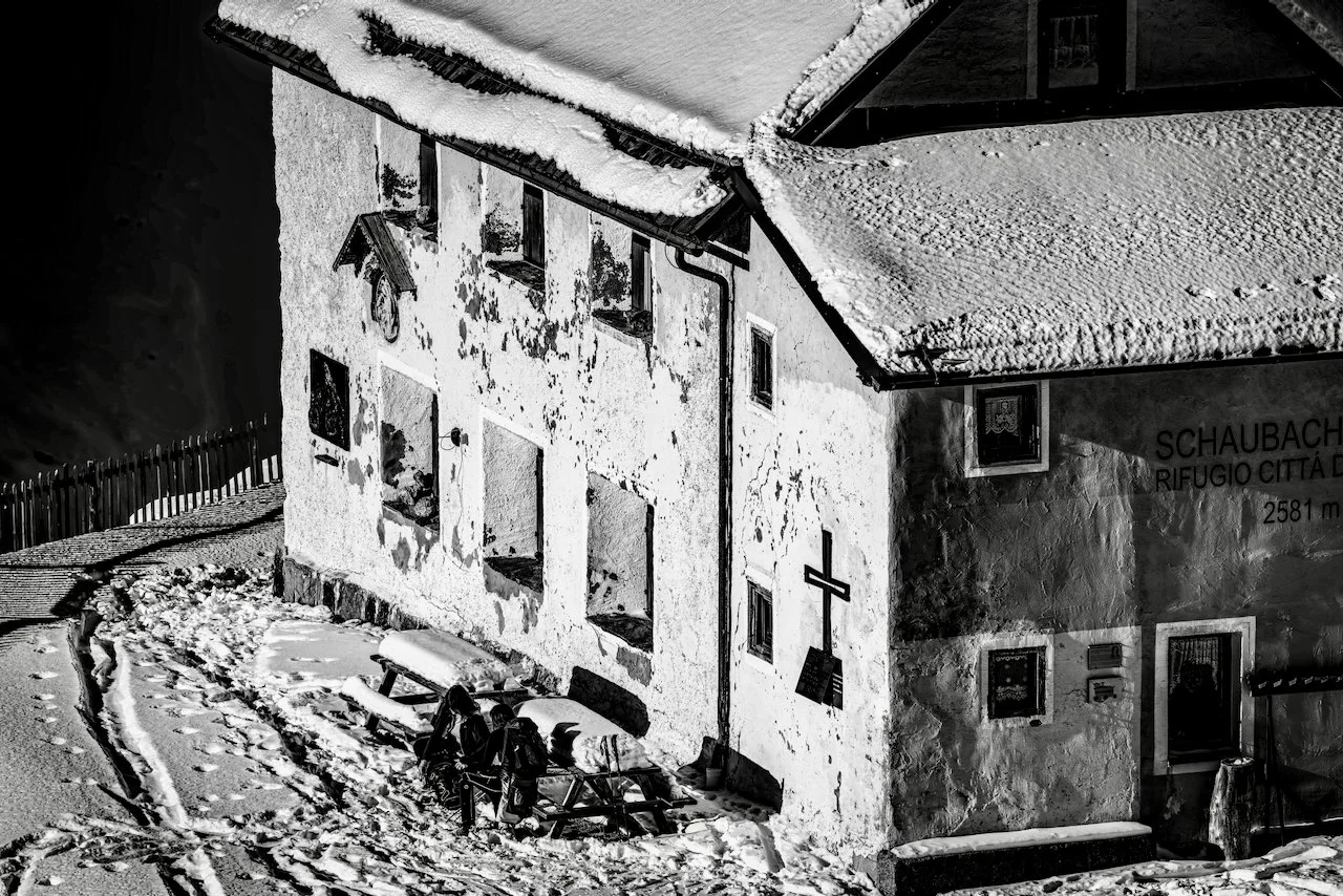 Black and white photo of a snow-covered mountain refuge building named 'Schaubach' with a cross on the exterior wall, snow on the roof, and a small group of hikers with backpacks sitting at a wooden picnic table outside on a snowy trail.