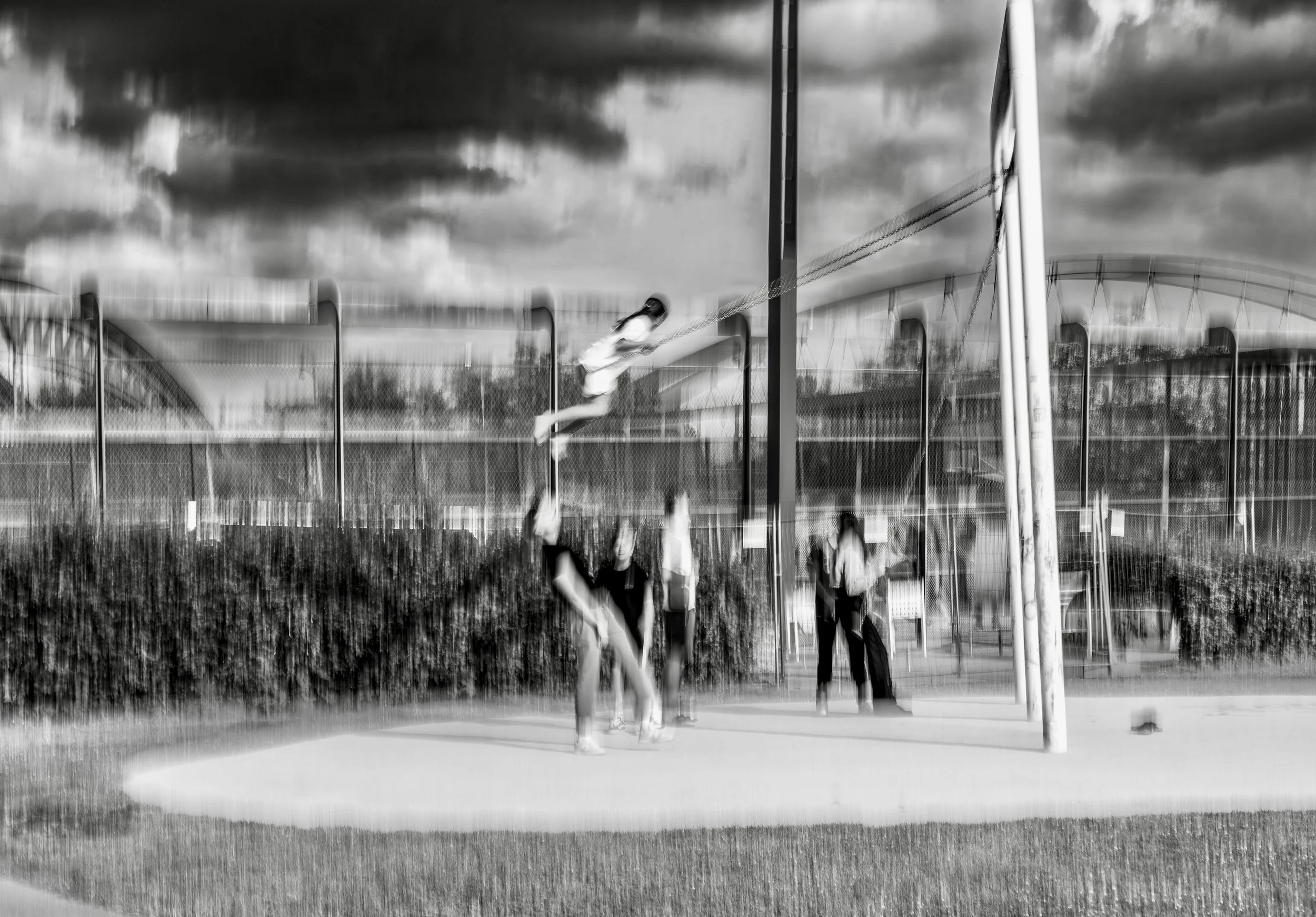 Black-and-white photo of a girl on a swing at a playground, with three other children standing nearby. A bridge and cloudy sky are visible in the background.