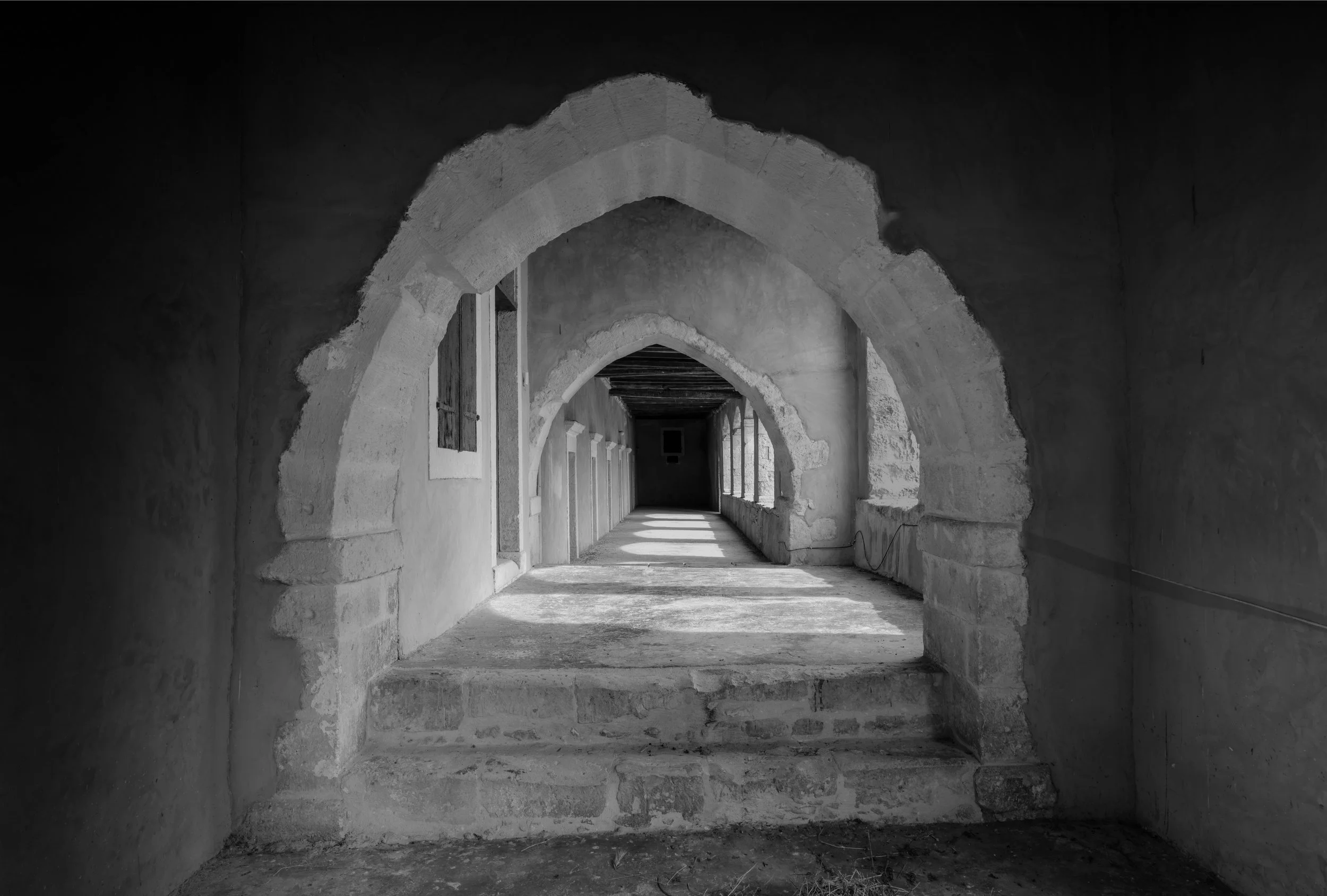 Black and white photo of a corridor with arched doorways, stone walls, and a window at the far end.