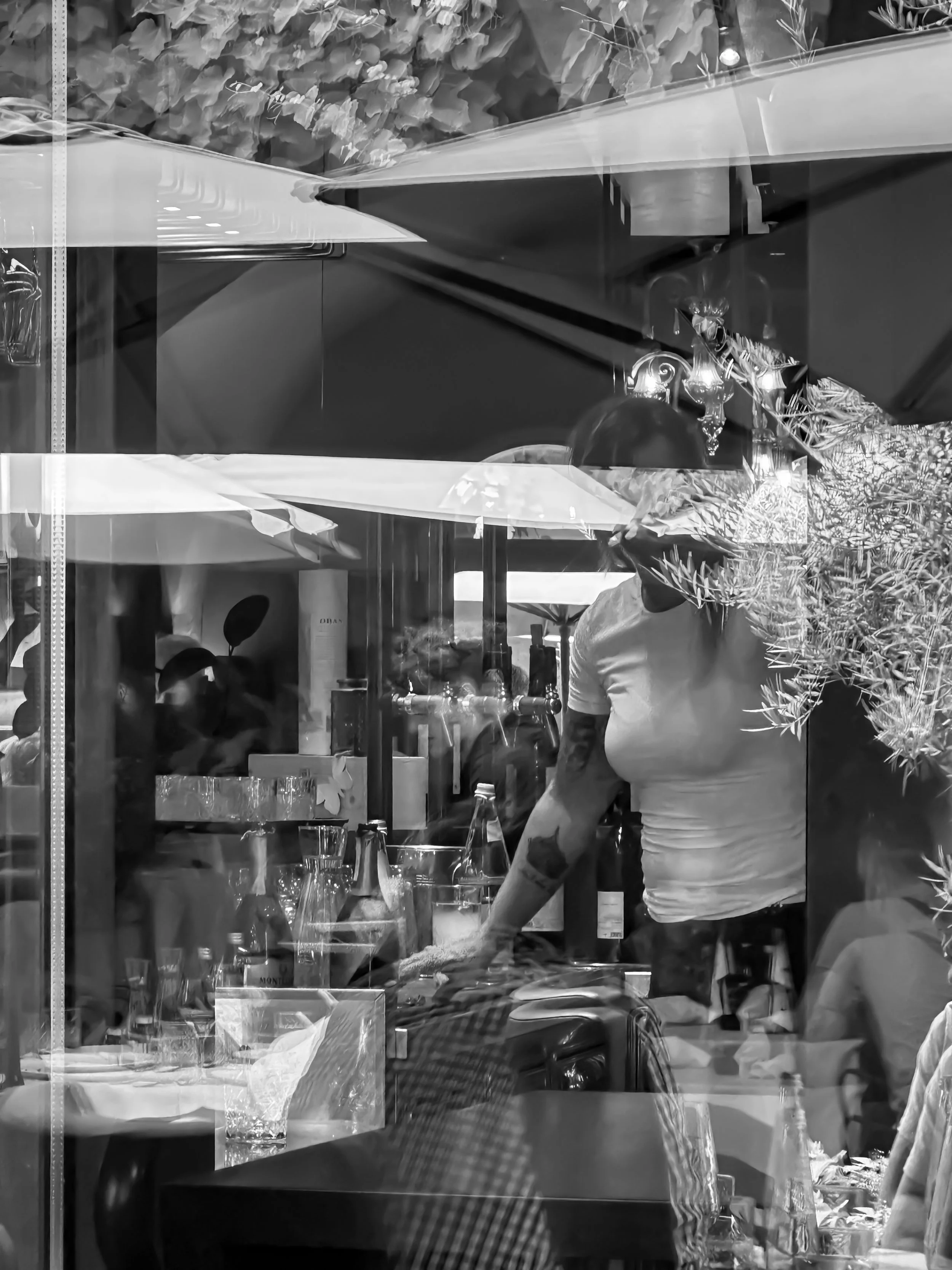 Reflective glass window with a view of an outdoor scene with trees, and a man with a beard and tattoos wearing a white T-shirt standing behind a table set with glasses and bottles, inside a restaurant or cafe.