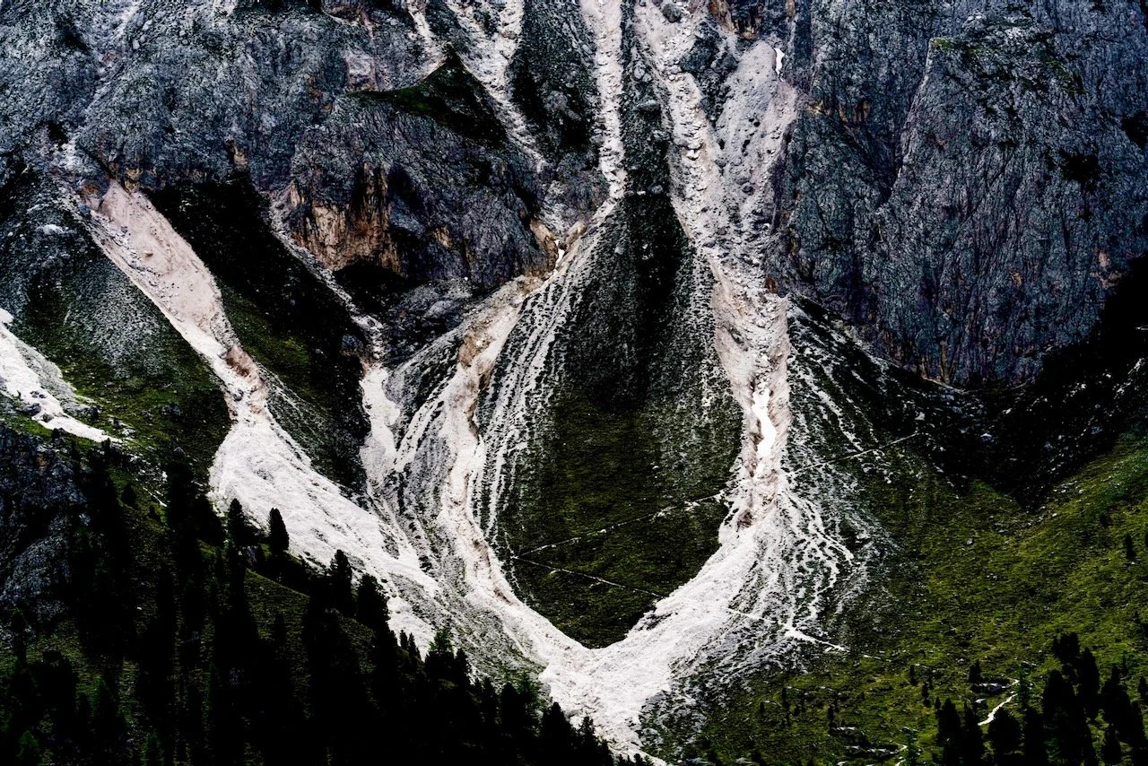 A mountain landscape with steep, rocky slopes, patches of snow, and a lush green forest at the base.