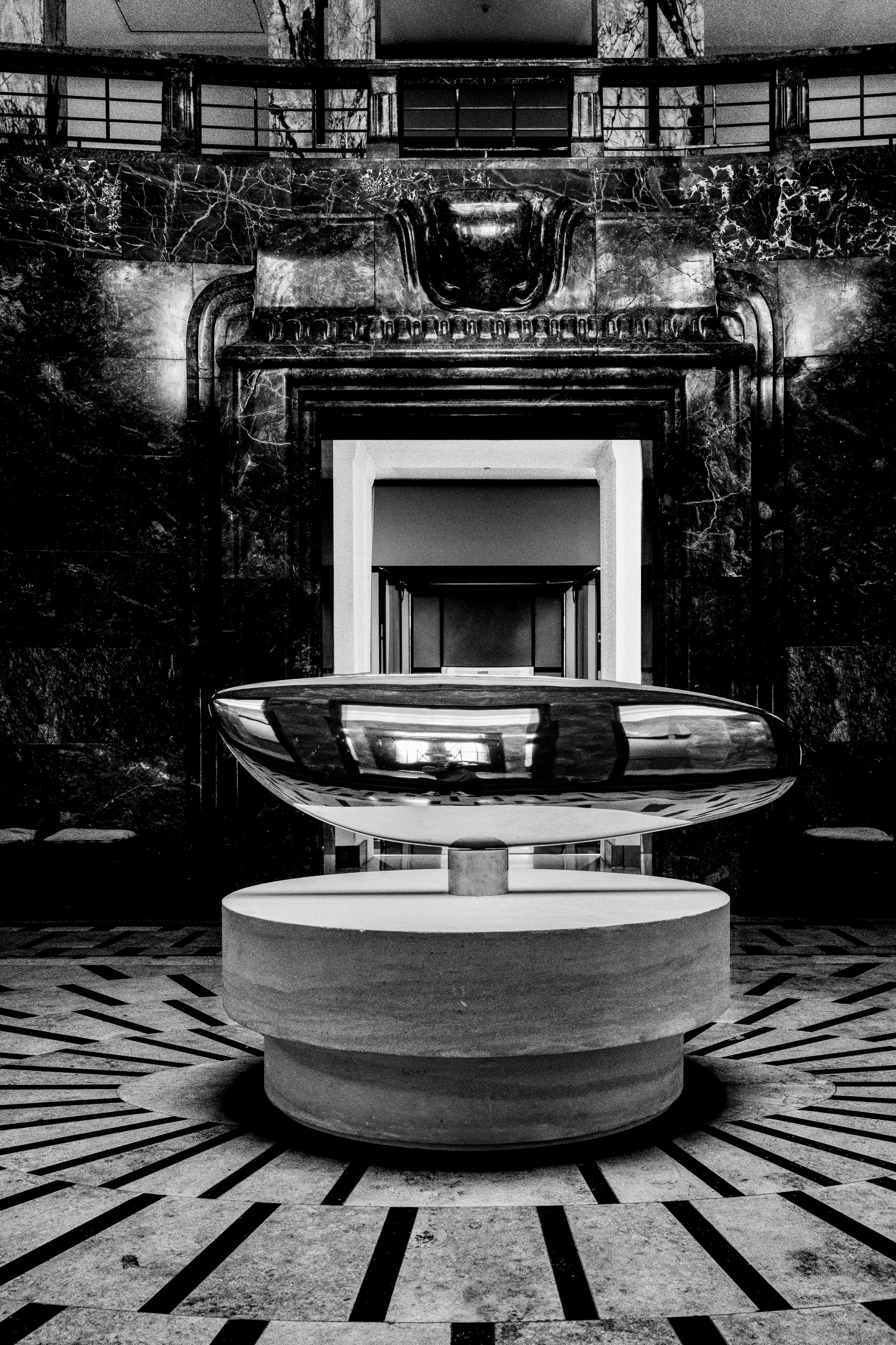 Black and white interior featuring a modern sculpture of a boat on a circular pedestal, with a marble wall and a staircase and walkway in the background.