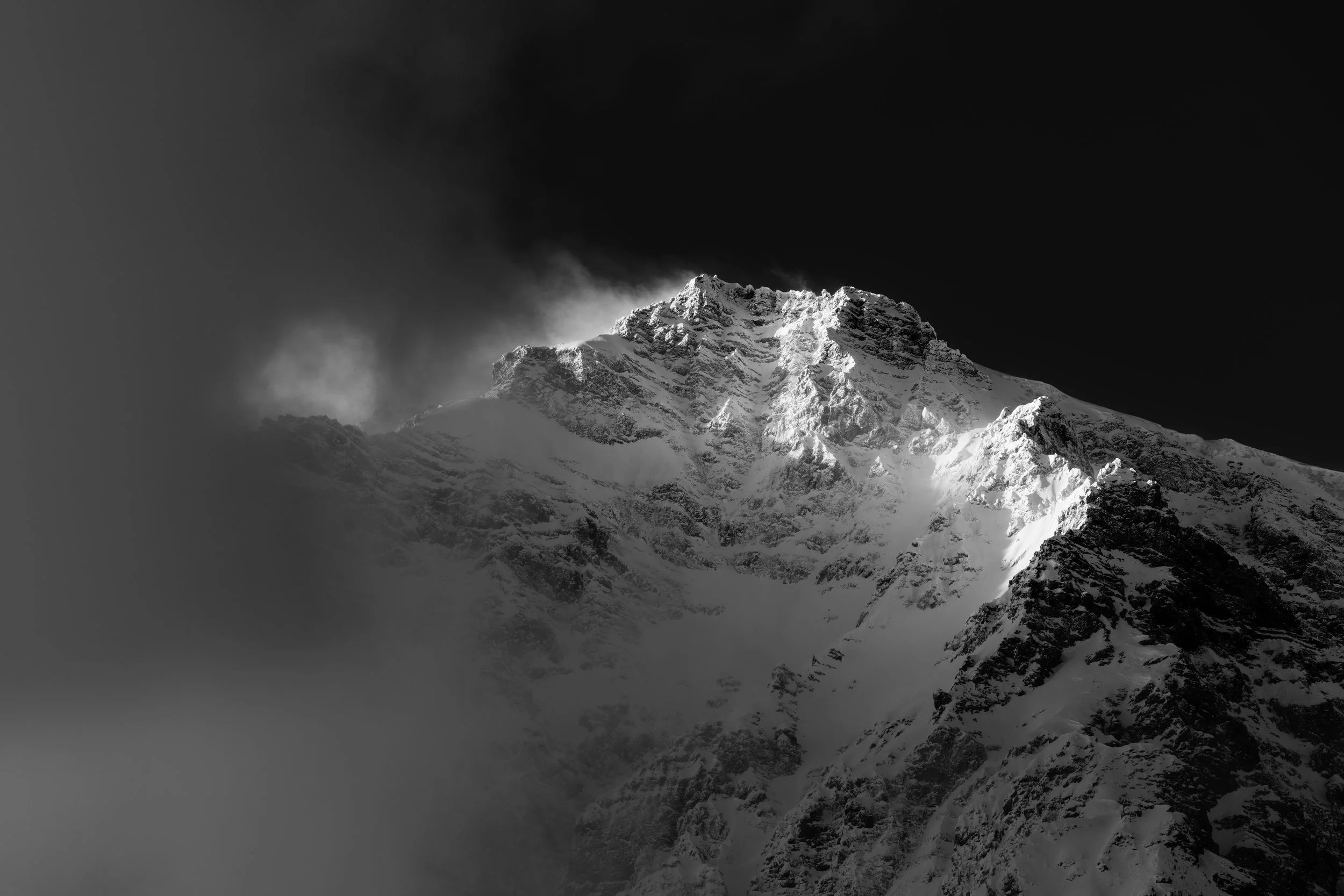 Black and white photo of a snow-covered mountain peak against a dark sky with mist or clouds at the base.