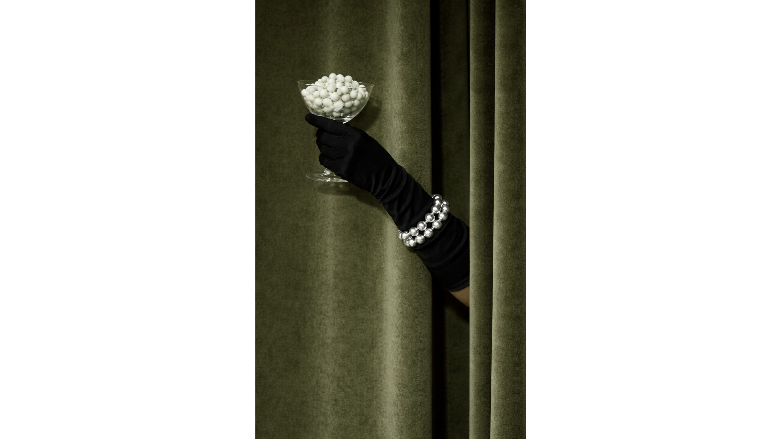 A hand wearing a black glove with pearl bracelets holding a glass bowl filled with white balls against a dark green curtain background.