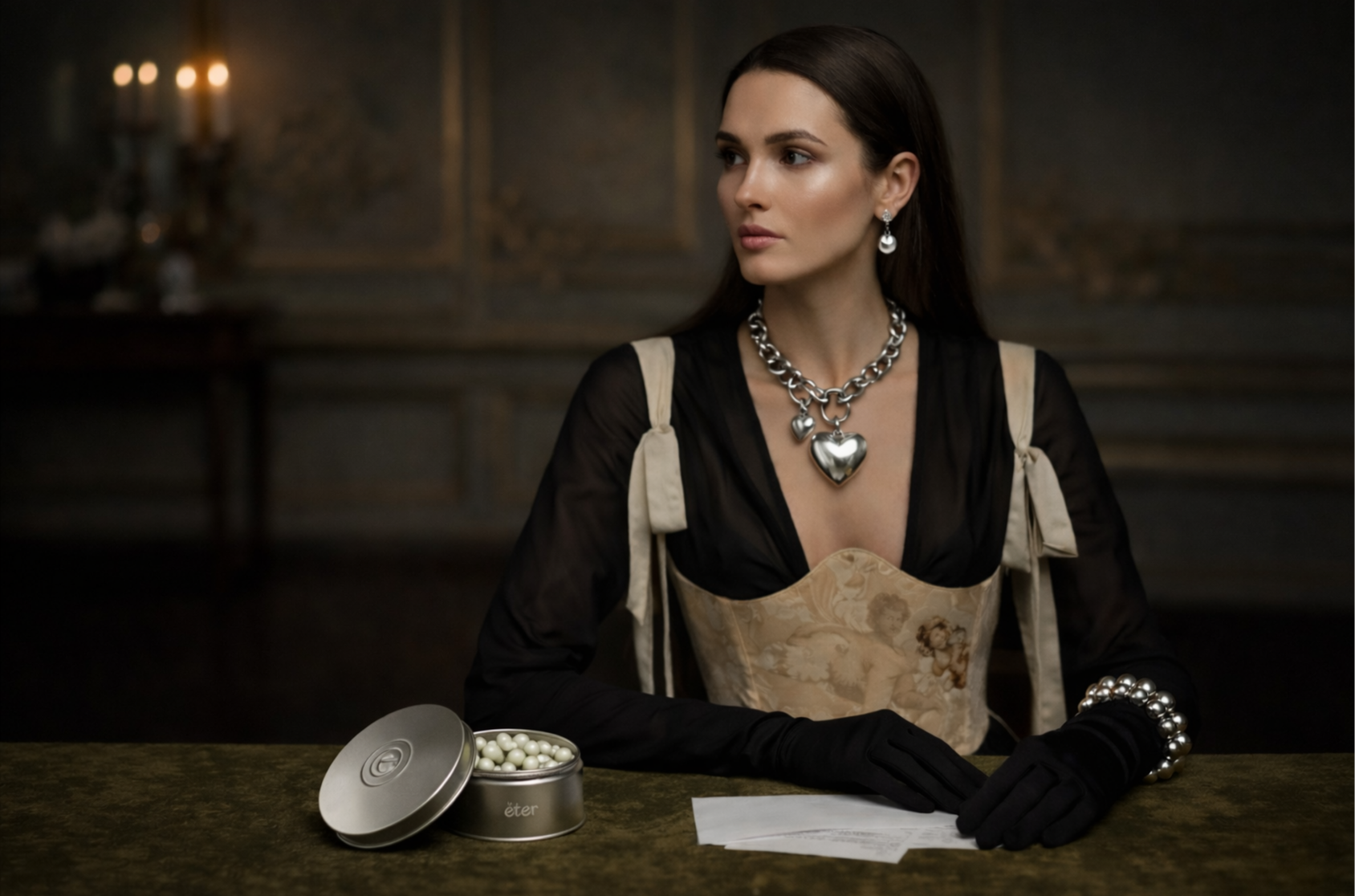 A woman with brown hair is sitting at a table, wearing a black sheer top, a beige patterned dress, large silver necklace with a heart pendant, matching earrings, and a pearl bracelet. There is a silver tin with a lid open to reveal white ball-shaped objects on the table in front of her, with a piece of paper and a half-filled glass of water nearby. The background is dimly lit with candles.