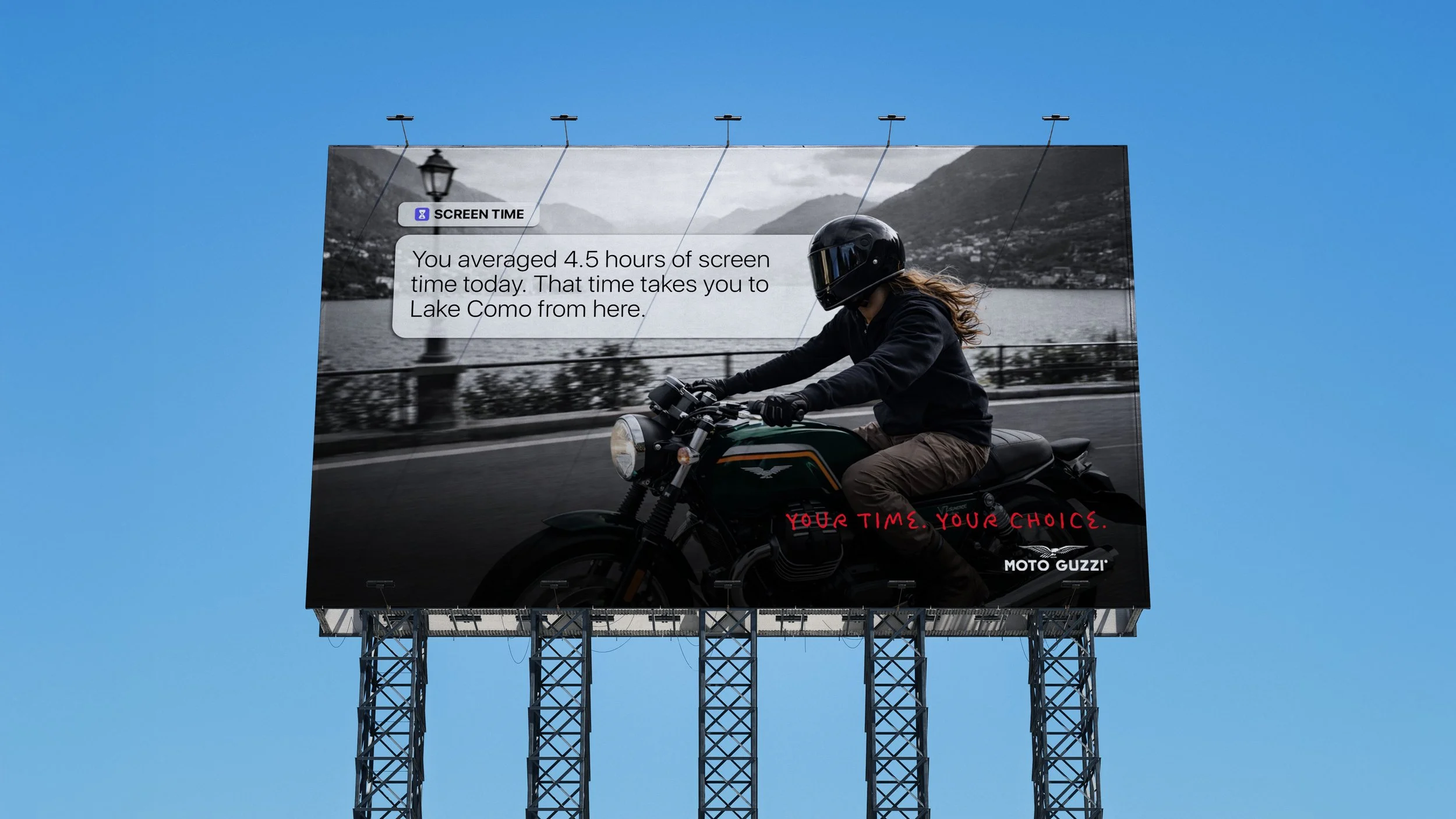 Billboard showing a person riding a motorcycle beside a lake, wearing a helmet, with mountains in the background. The billboard includes a message about screen time and travel distance, with the words "YOUR TIME. YOUR CHOICE." and the Moto Guzzi logo.