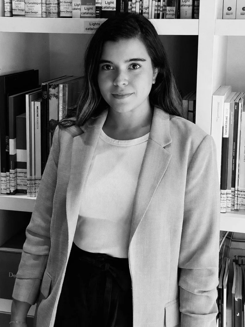 A woman with long dark hair, wearing a blazer and a light-colored shirt, standing in front of bookshelves in a library.