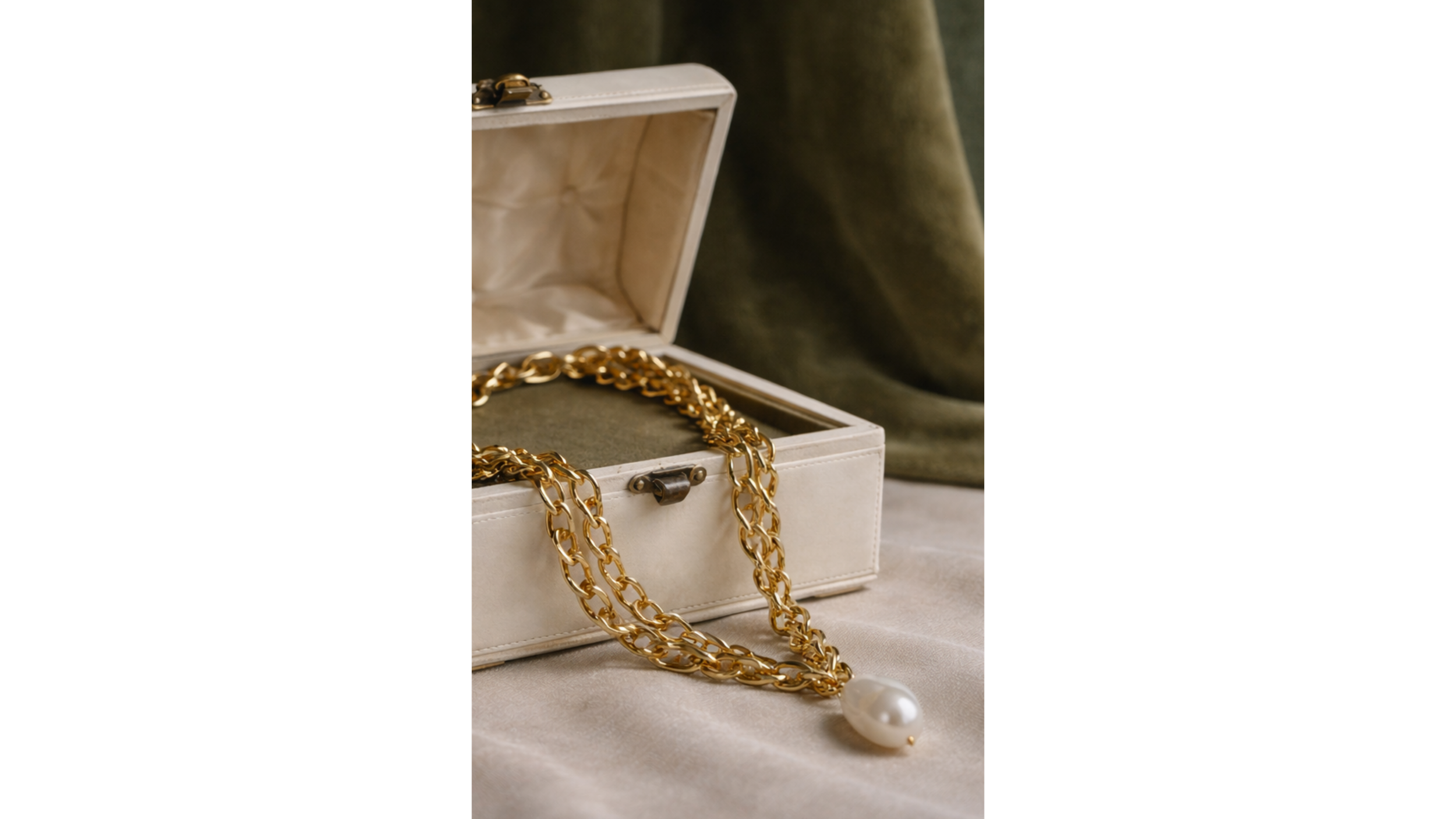 A white jewelry box with gold chains and a pearl necklace inside, placed on a soft fabric surface.