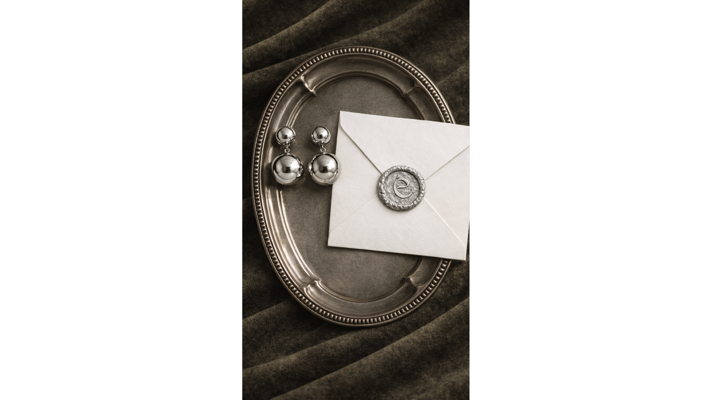 Silver jewelry, including a pair of silver ball earrings, displayed on a silver tray with an envelope sealed with a wax stamp featuring an 'e' in cursive, all set on a dark textured fabric background.