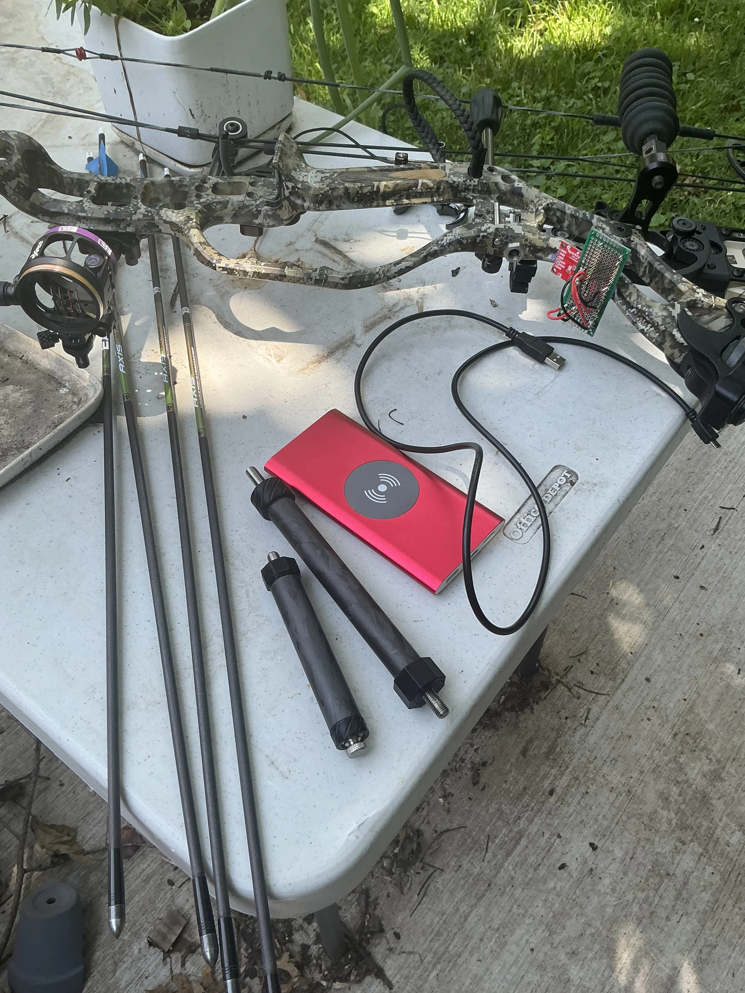 A compound bow with sensor package installed to measure vibration and energy. IMU, power bank, and bow stabilizers visible with custom arrows on the table.